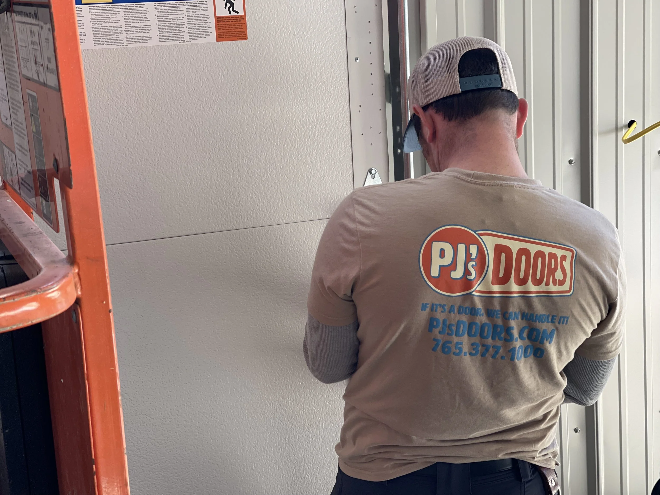 Two workers on a yellow scissor lift installing drywall on an overhead ceiling of a garage. One worker is wearing a black helmet, and the other is wearing glasses and a blue jacket. The garage door is open with a view of a parking lot and neighboring buildings.