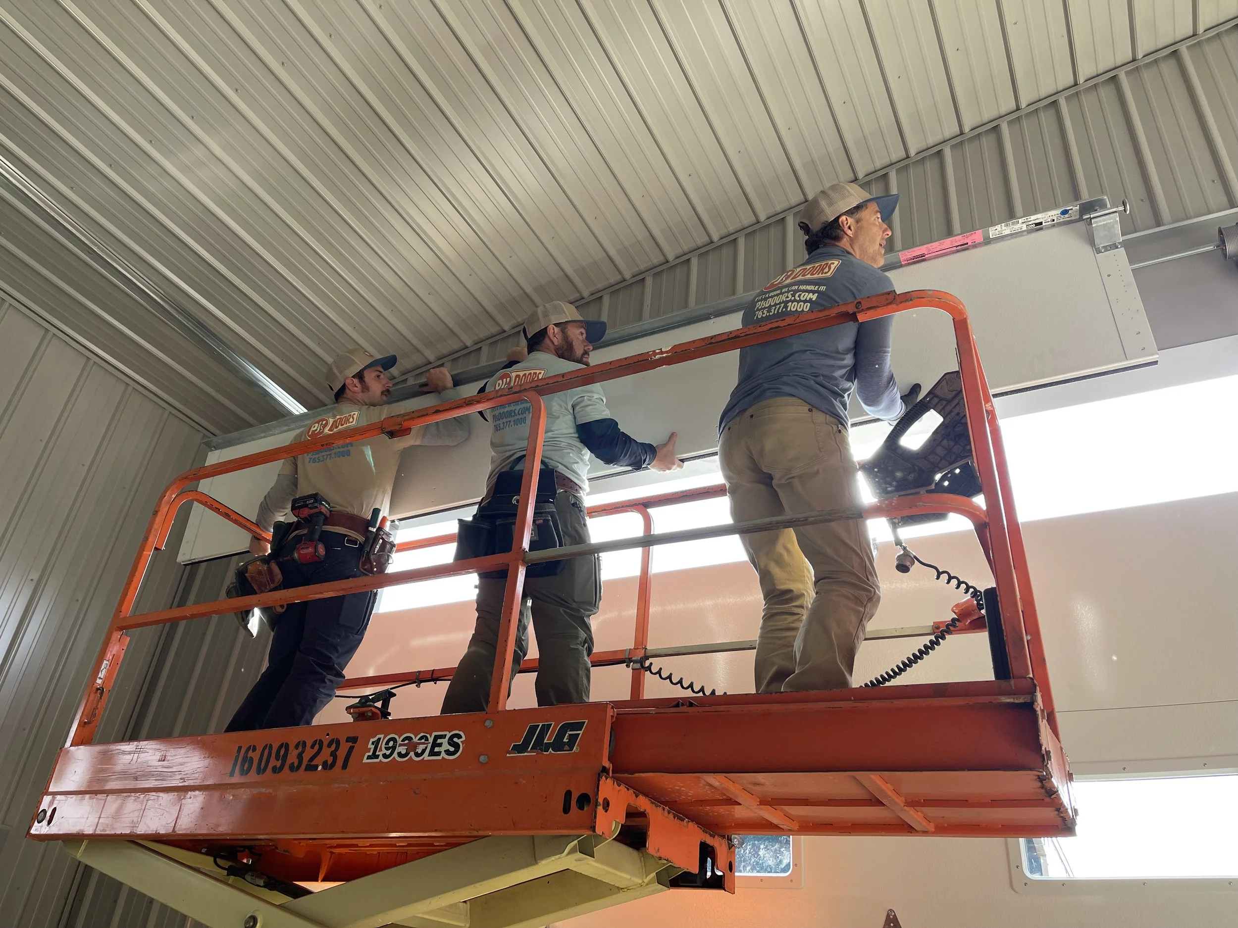 Two workers on a yellow scissor lift installing drywall on an overhead ceiling of a garage. One worker is wearing a black helmet, and the other is wearing glasses and a blue jacket. The garage door is open with a view of a parking lot and neighboring buildings.