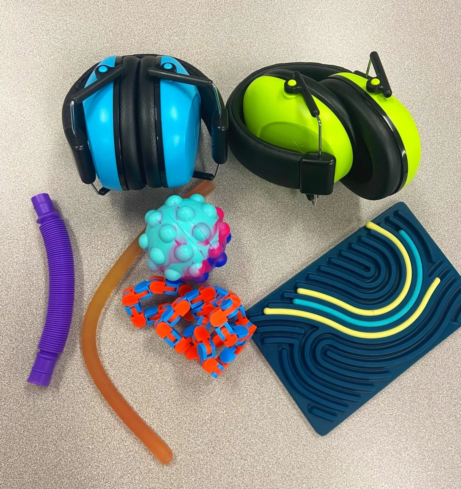 Two pairs of colorful noise-canceling headphones, one blue and the other green, placed on a gray surface. Nearby are various flexible tubes and textured objects, including a purple, brown, and orange tube, a multicolored spiky ball, a red and blue chain-like object, and a blue foam pad with yellow wavy lines.
