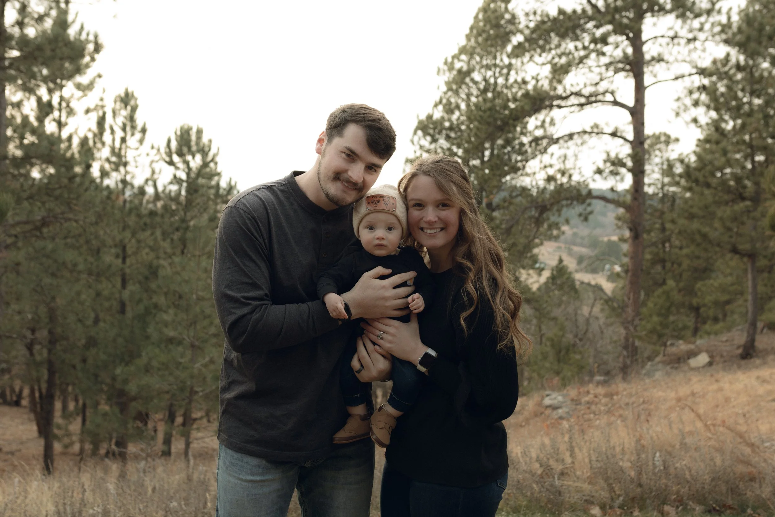 A family of three standing outdoors in a wooded area, smiling at the camera. The man has short dark hair and wears a dark long-sleeve shirt, holding a baby. The woman has long wavy brown hair and wears a black top. The baby, who has light-colored hair, wears a beige hat and dark clothing.