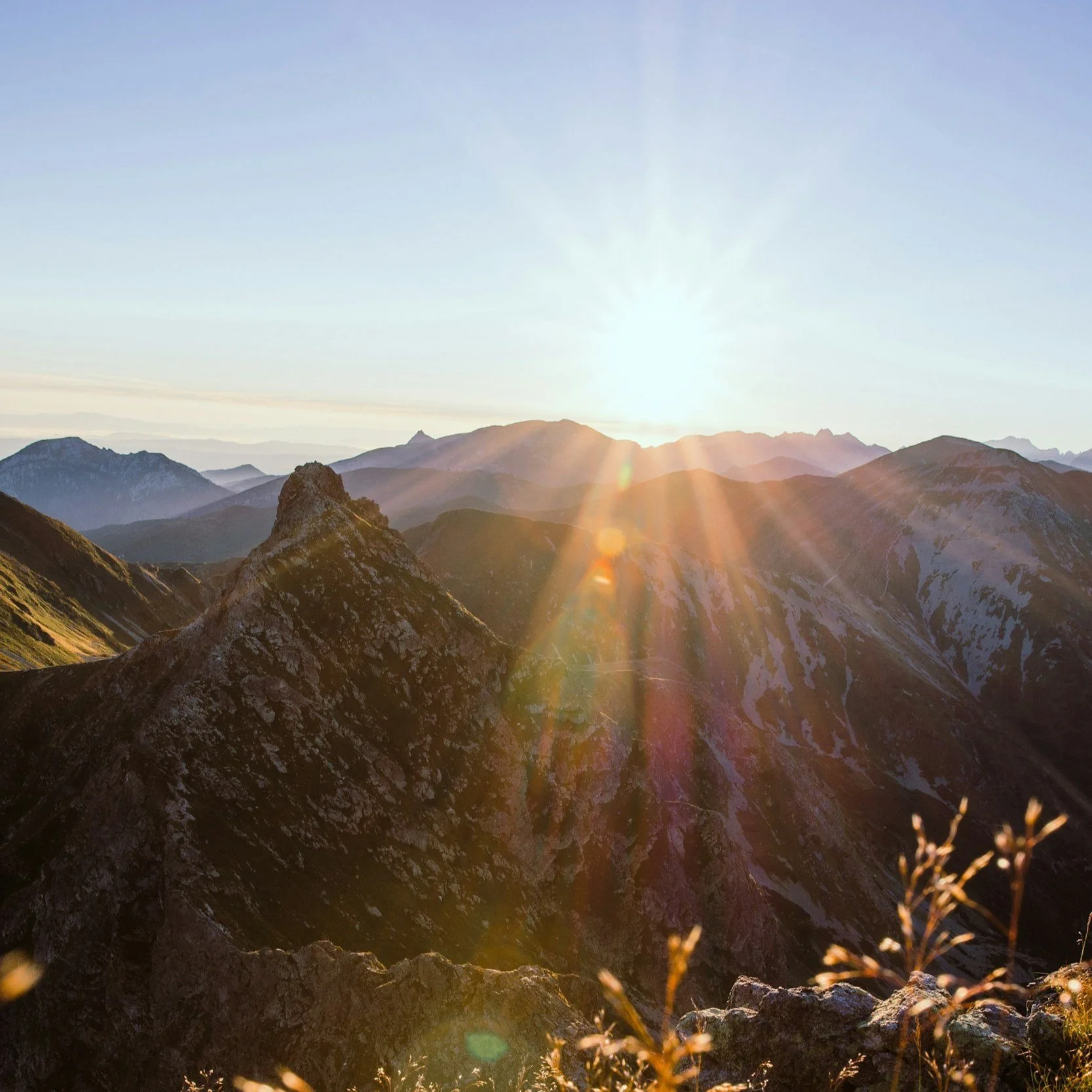 Sunrise over mountain range with sun rays illuminating rocky peaks and a clear sky.