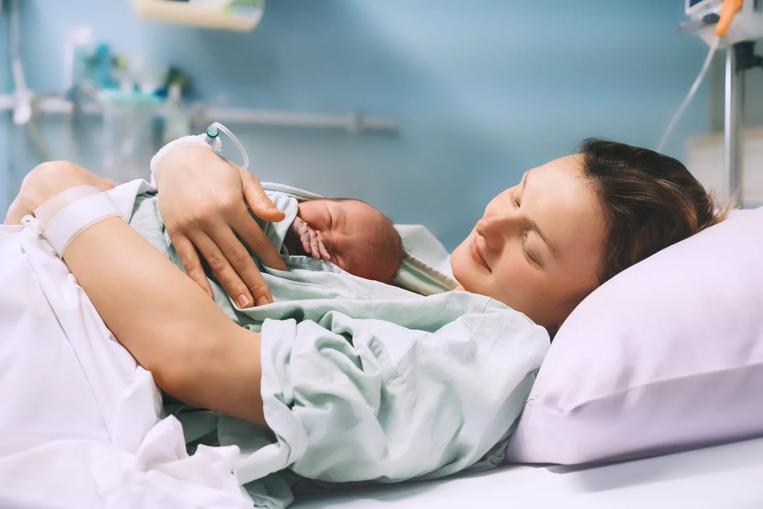 A woman lying on a hospital bed holding a newborn baby close to her chest, with medical equipment visible in the background.