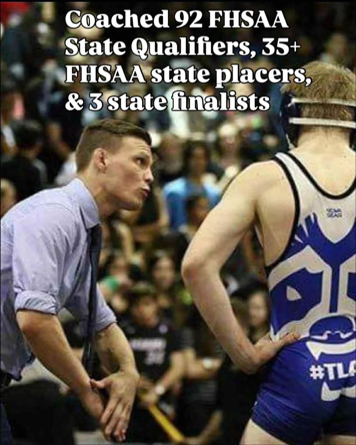 A coach with a focused expression coaching a wrestler during a match at a wrestling event with a large crowd in the background.