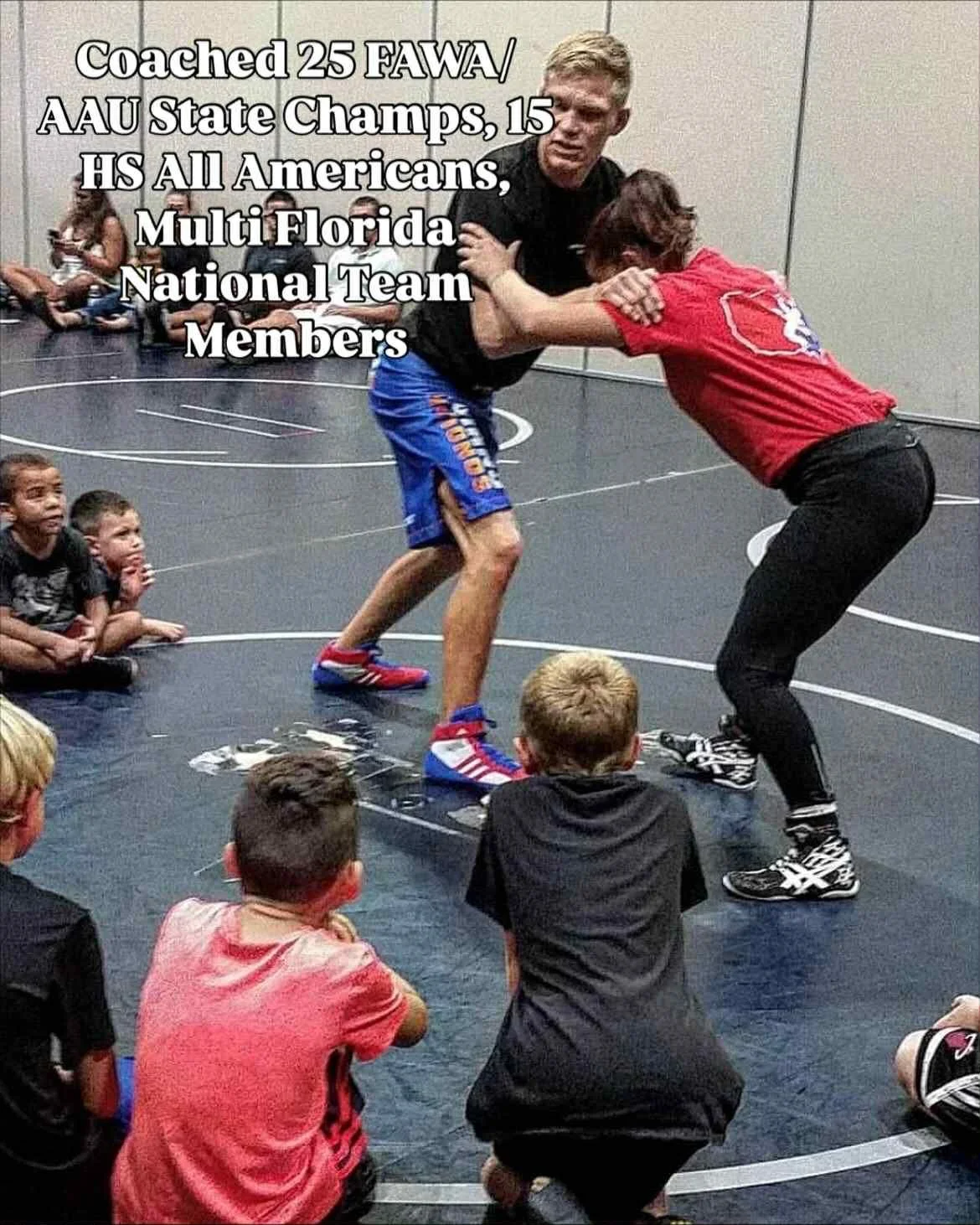 A wrestling or grappling class for children in progress, with kids sitting on the floor watching two adults wrestle on a mat.
