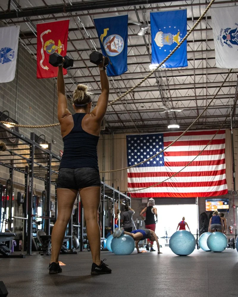 Woman lifting dumbbells in a gym with American flag and military flags hanging from the ceiling.