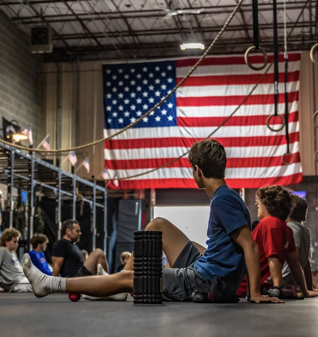 Children sitting on the floor in a gymnasium with an American flag and gymnastic rings in the background.