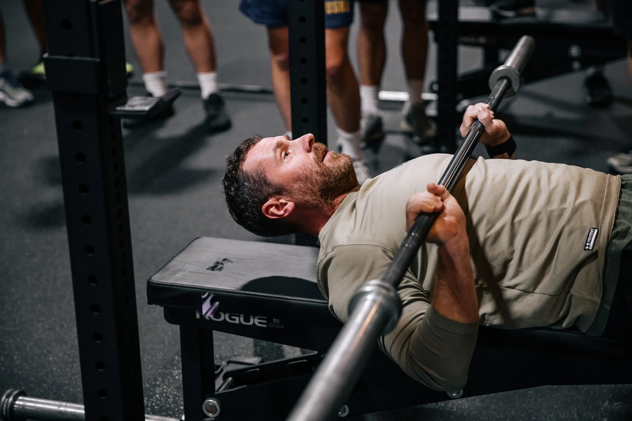 A man lying on a bench at a gym lifting a barbell during a workout session, with other people working out in the background.