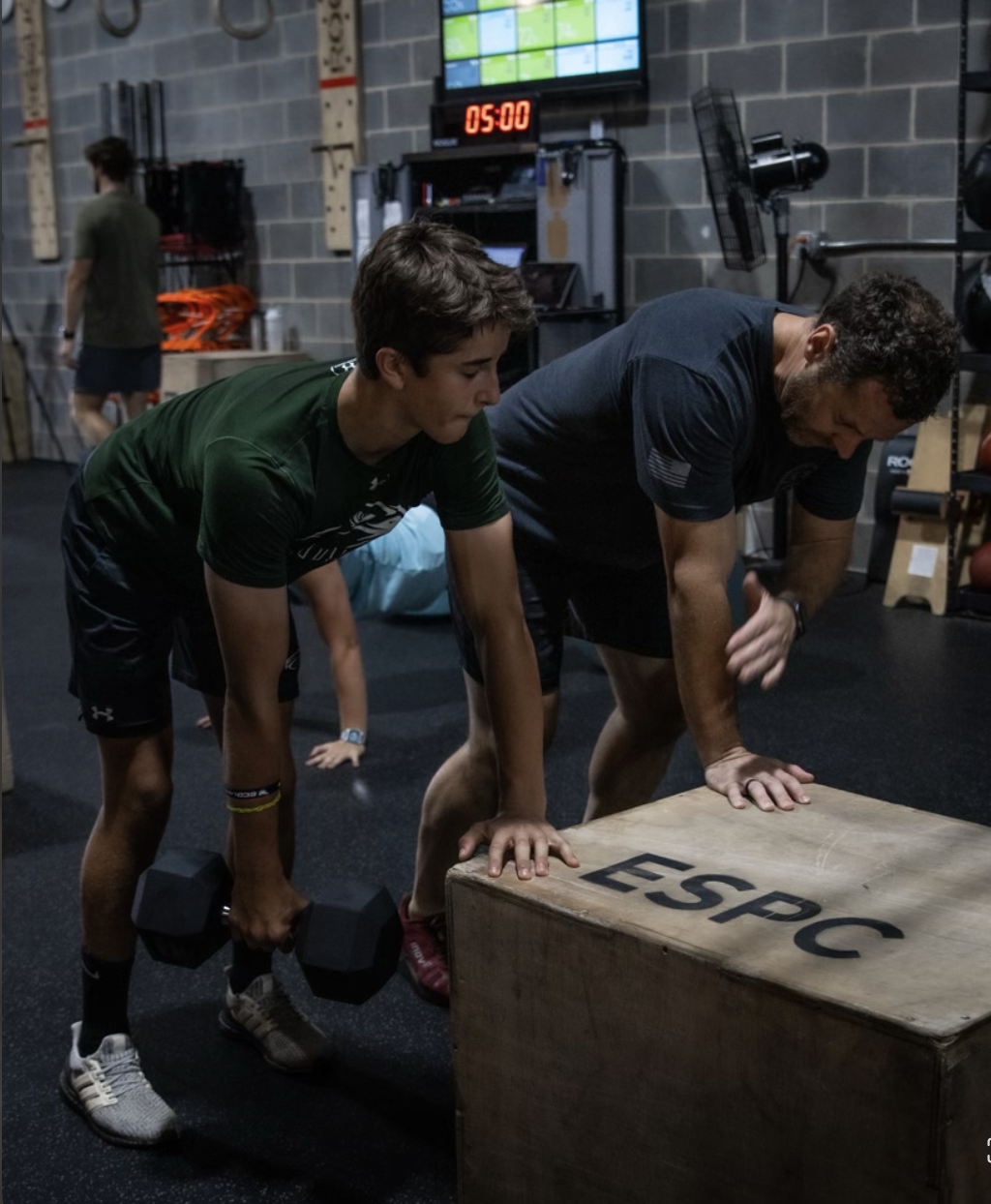 A young man and coach doing a plank exercise in a gym, with the young man holding dumbbells and the coach supporting him from the side.