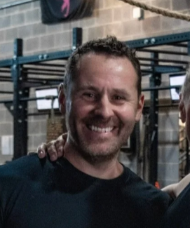 A smiling man with short curly hair and a beard, wearing a black shirt, standing in a gym with workout equipment in the background.