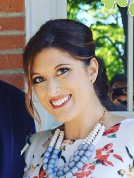 A woman with wavy brown hair smiling outdoors with trees and sunlight in the background.
