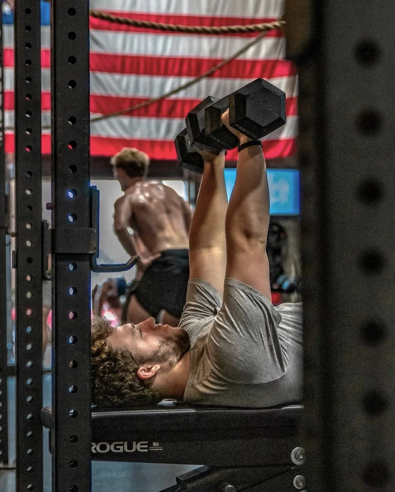 A man lying on a workout bench in a gym lifting dumbbells overhead, with an American flag hanging on the wall in the background.