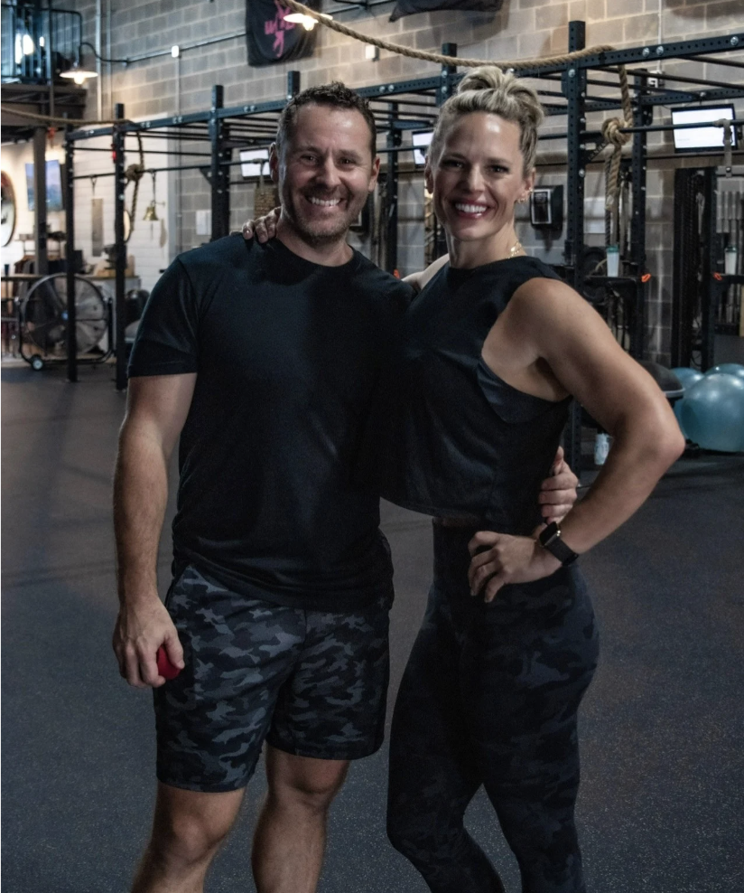 A man and woman smiling at the camera inside a gym, standing close with arms around each other. The gym has black equipment, gym balls, and climbing ropes.