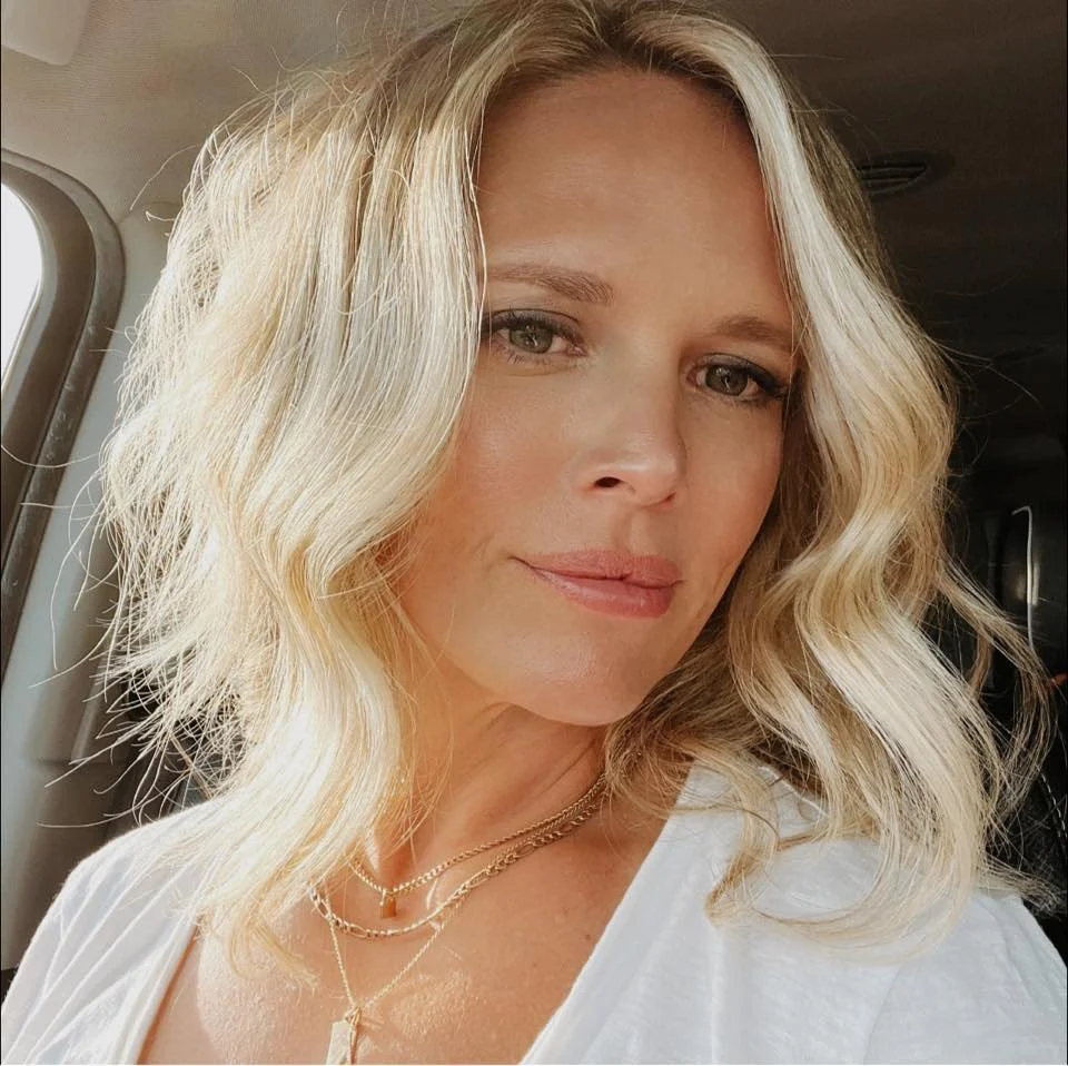 A woman with shoulder-length blonde hair, styled in loose waves, sitting in a vehicle with a light-colored shirt and layered gold necklaces.