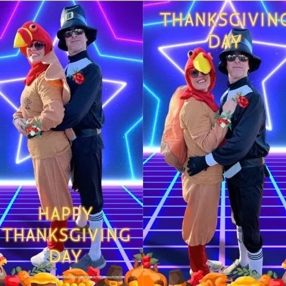 Two people are dressed in costumes, one as a turkey and the other as a pilgrim. They are standing together in front of a neon-lit background with geometric shapes and the words 'Happy Thanksgiving Day' and 'Thanksgiving Day' written on it. They are posing for the photo, hugging each other. The scene is festive with fall-themed decorations at the bottom.