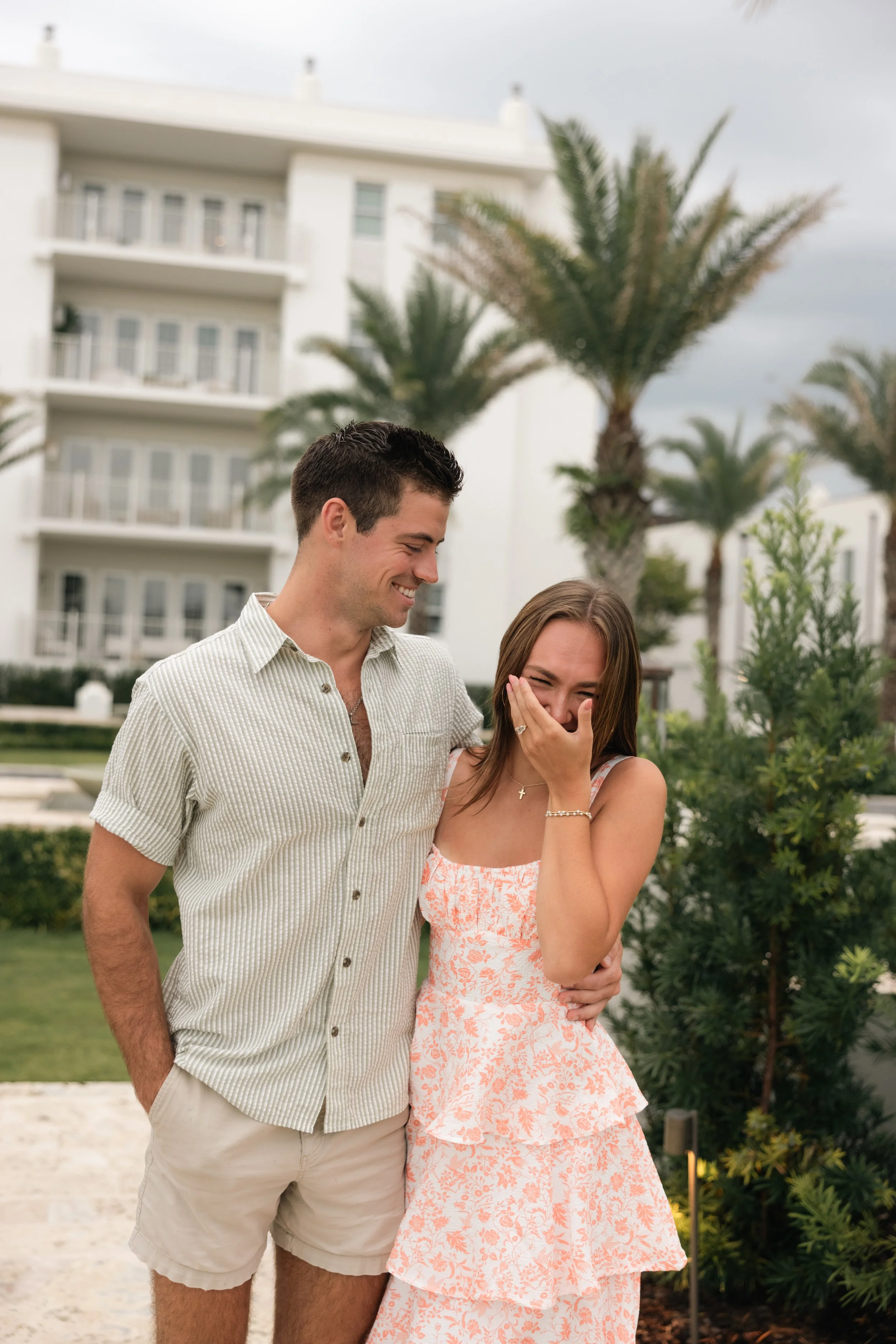 A young man and woman sharing a happy moment outdoors, with the man smiling and the woman covering her mouth in surprise, against a backdrop of palm trees and a modern white building.