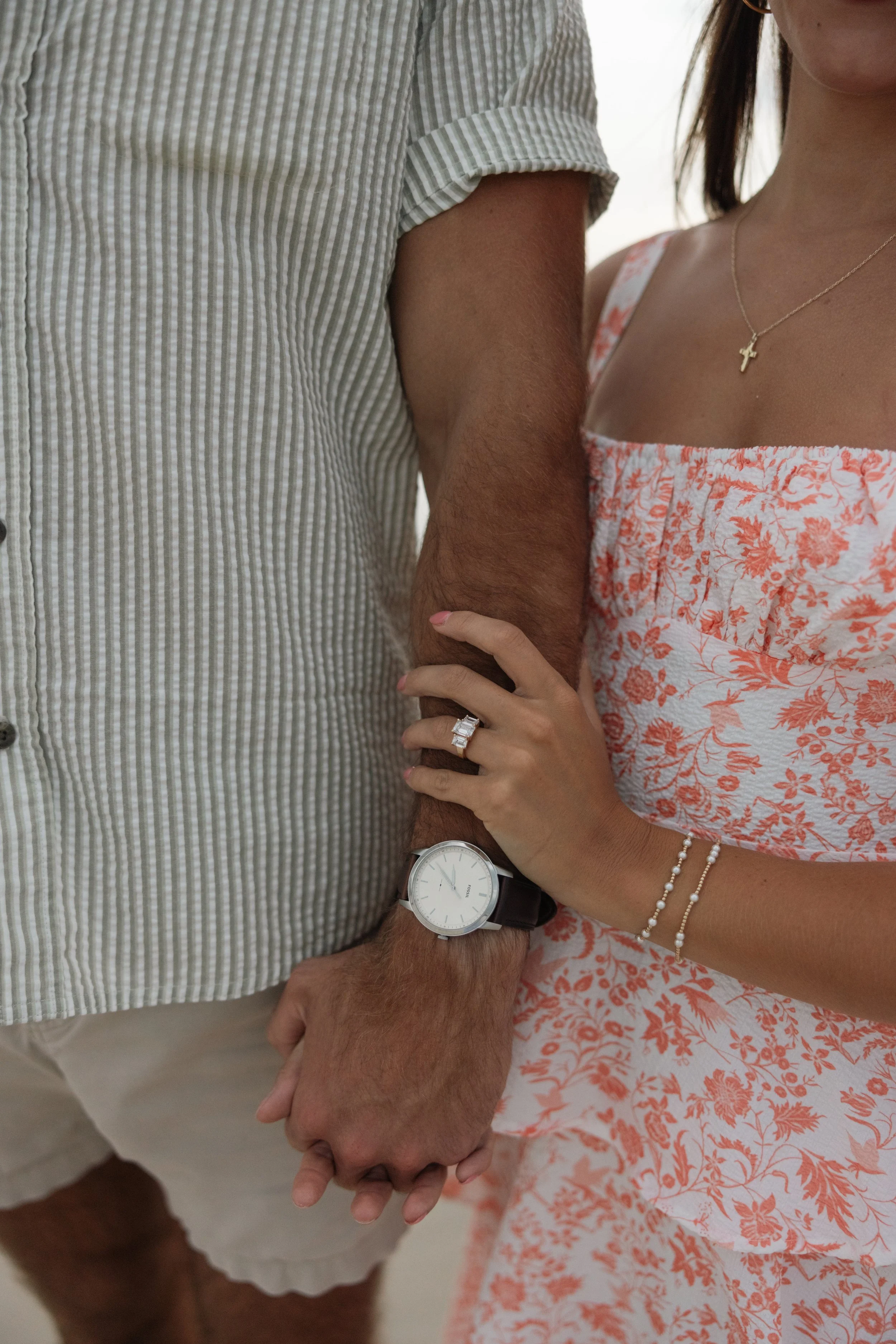 Close-up of a couple holding hands, showing their arms, with the woman wearing jewelry and the man wearing a watch.