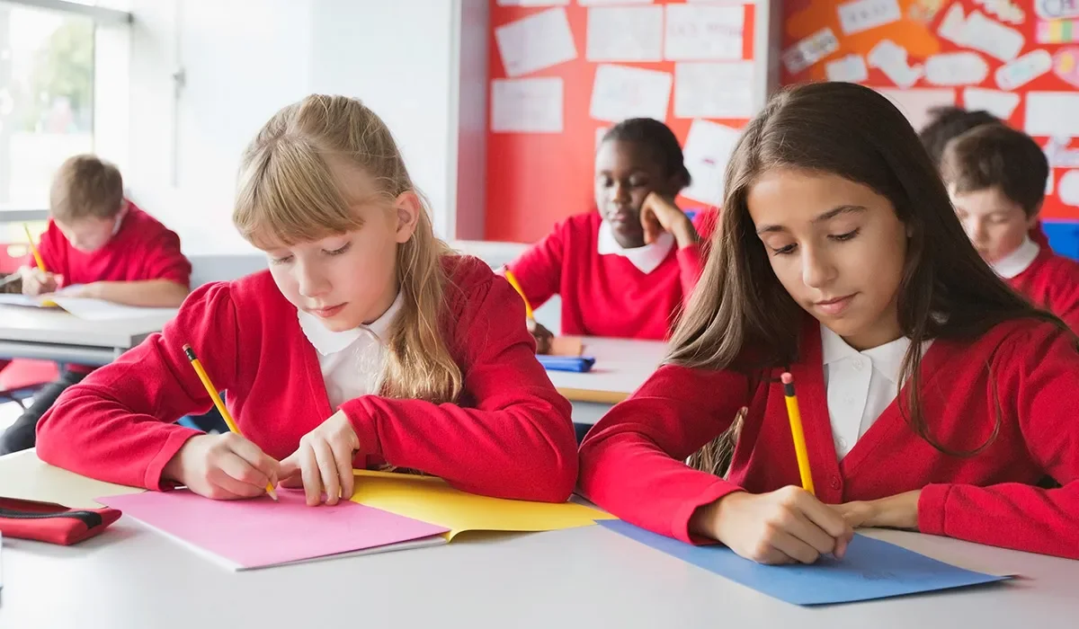 Young students in a classroom, wearing red sweaters, working on assignments at their desks with colorful paperwork.