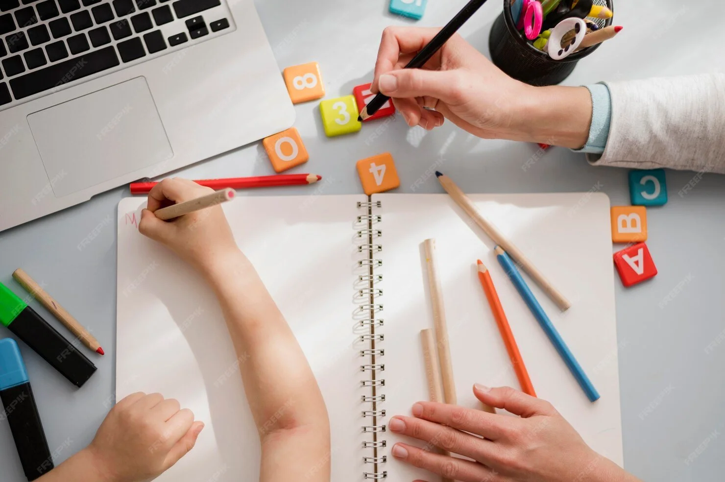 Two people working on a planning or learning activity at a desk with colored markers, foam blocks with numbers, Pokemon-themed tokens, and an open notebook.