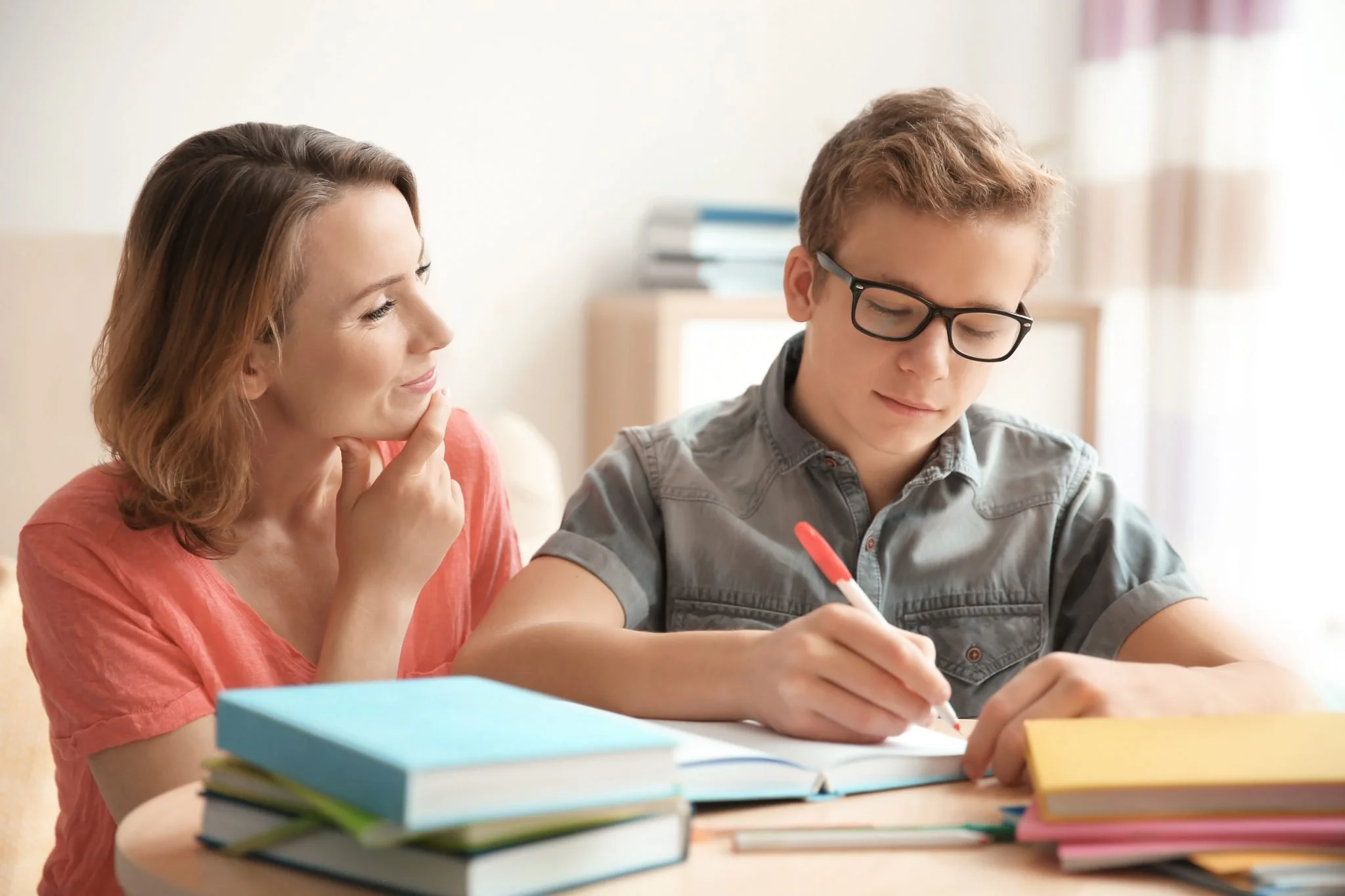 A woman looks at a teenage boy who is writing in a notebook at a table with stacked books.