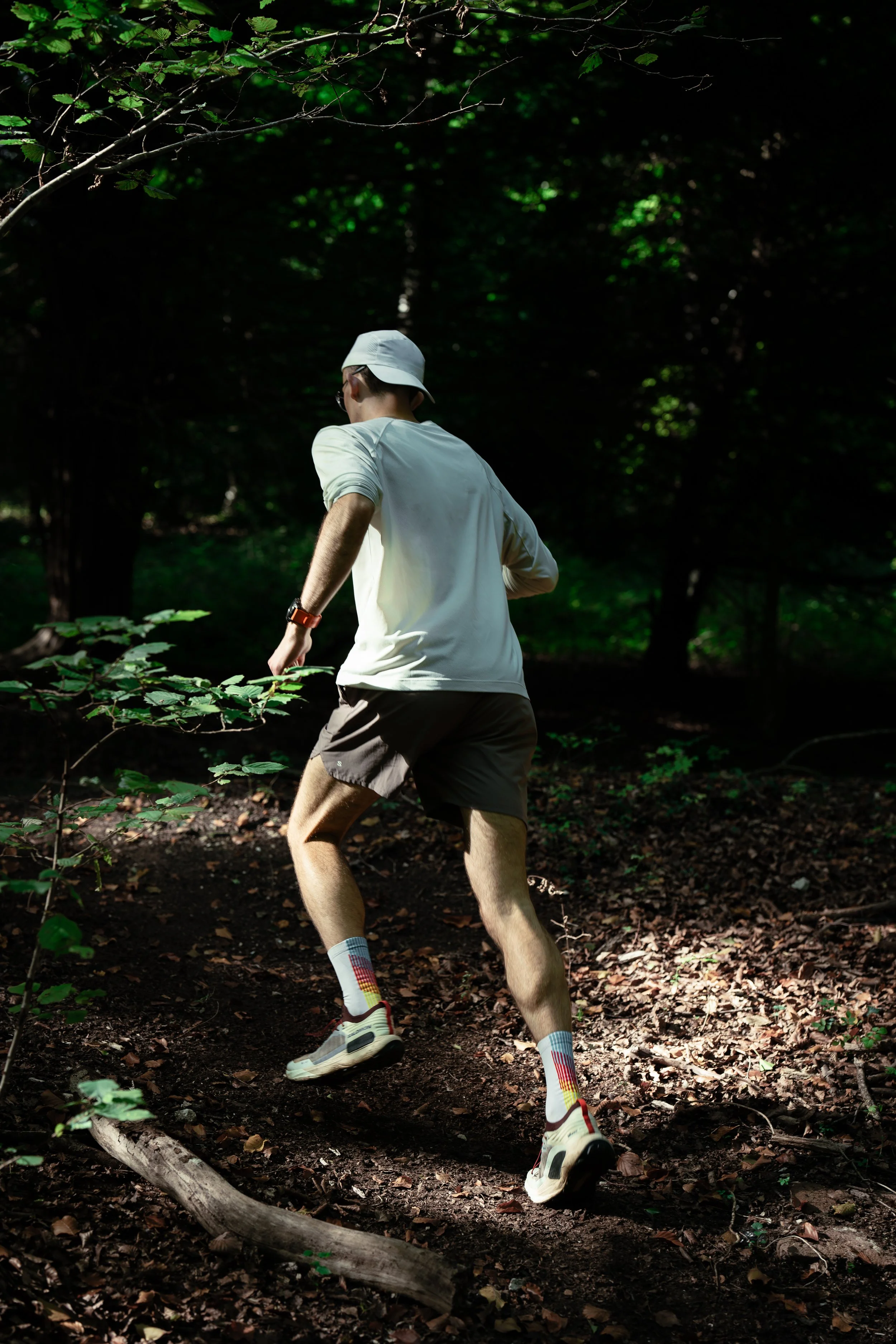 A man trail running through a wooded forest, wearing a white long-sleeve shirt, beige shorts, colorful patterned socks, and white running shoes, with sunlight filtering through the trees.