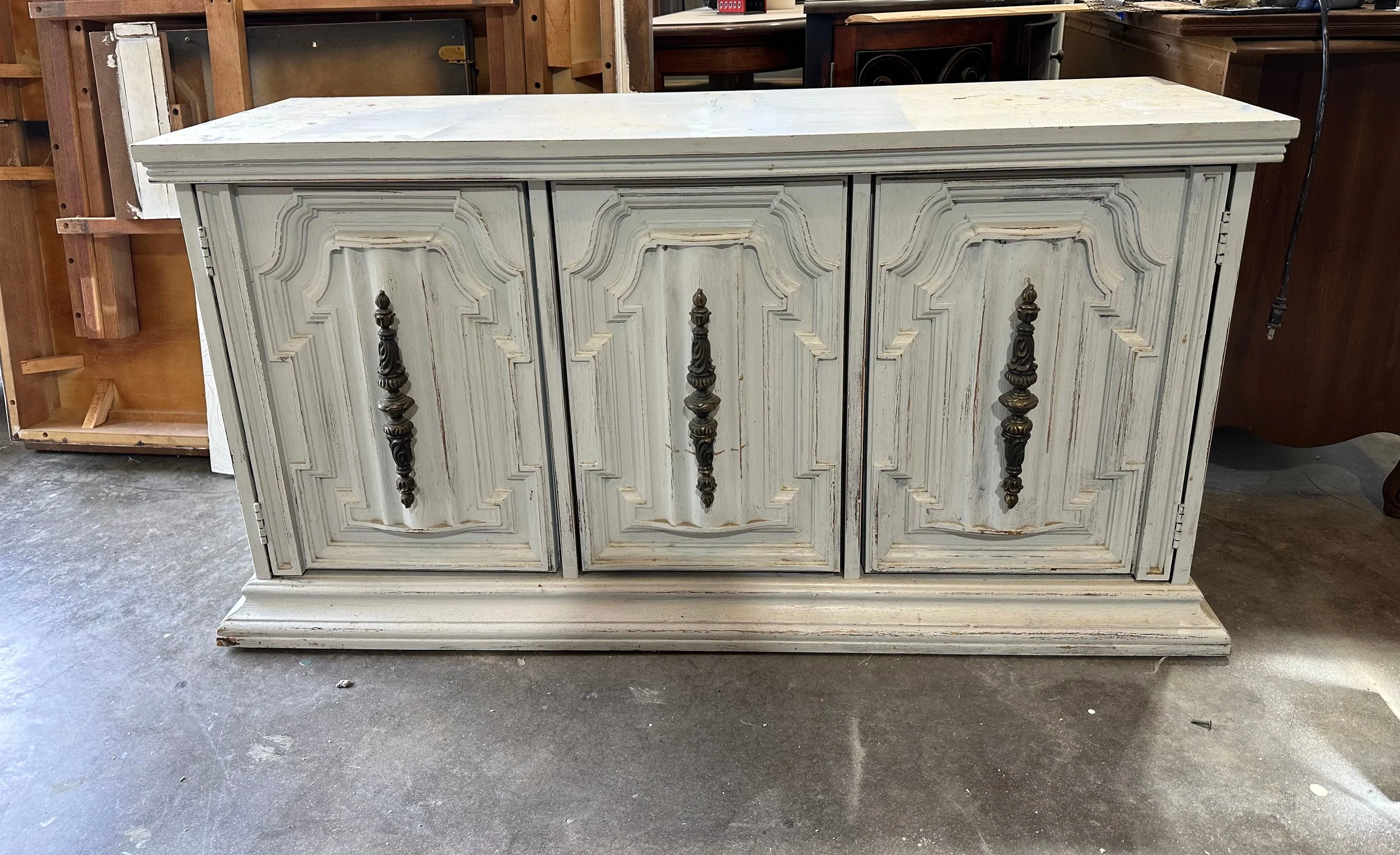 Vintage distressed white wooden cabinet with three doors and dark ornate handles, standing on a concrete floor in a workshop or storage space.