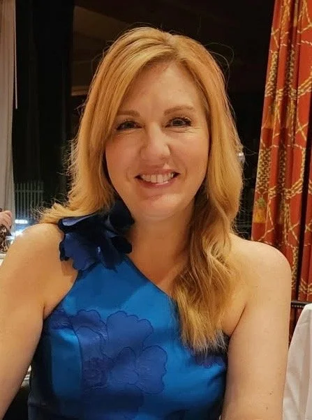 Trina Ellis smiling at a dinner table, wearing a blue dress with ruffled shoulder details, in a room with red curtains.