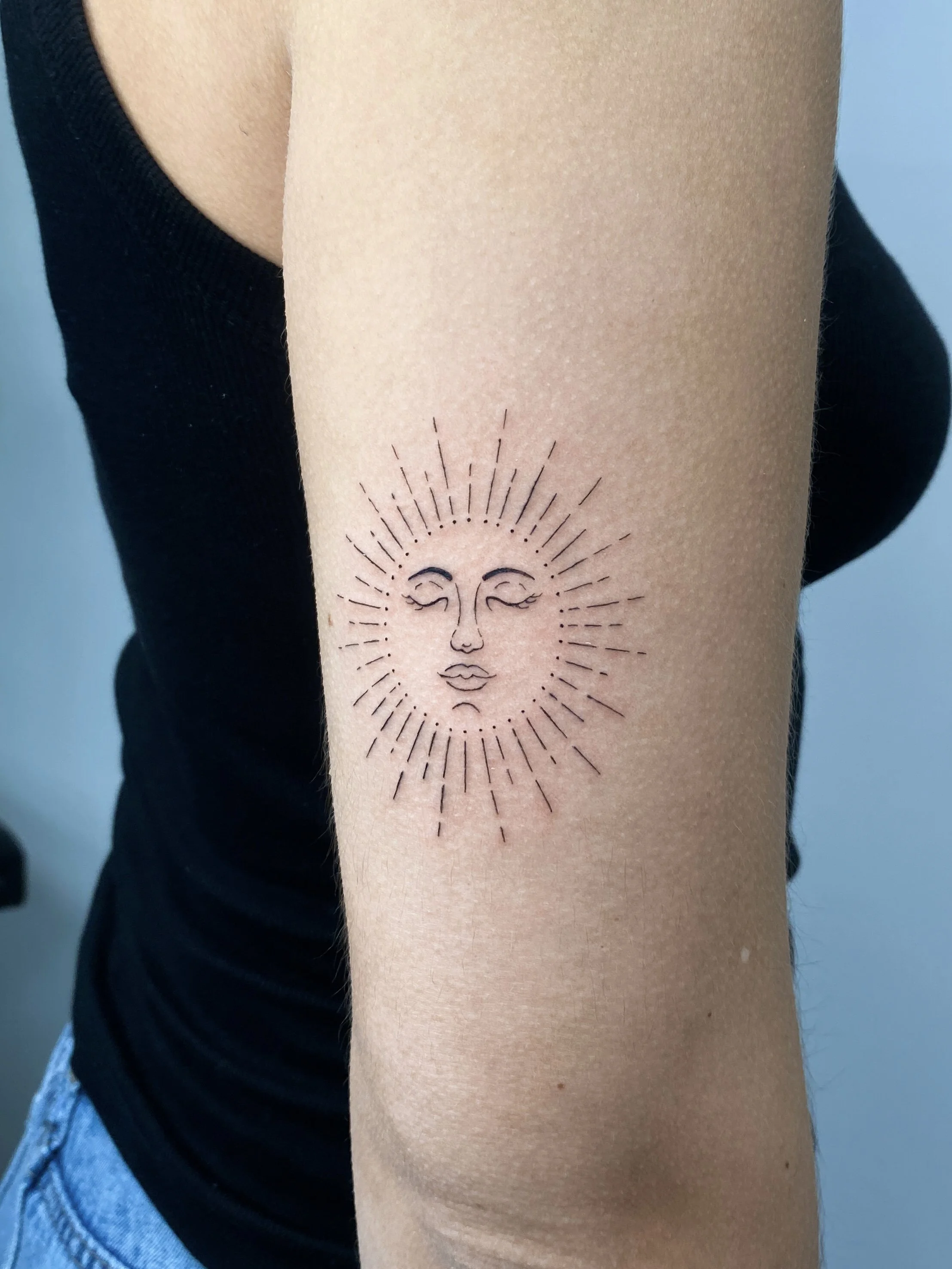 A tattoo of a smiling sun with a face, surrounded by radiating lines, on a person's forearm.