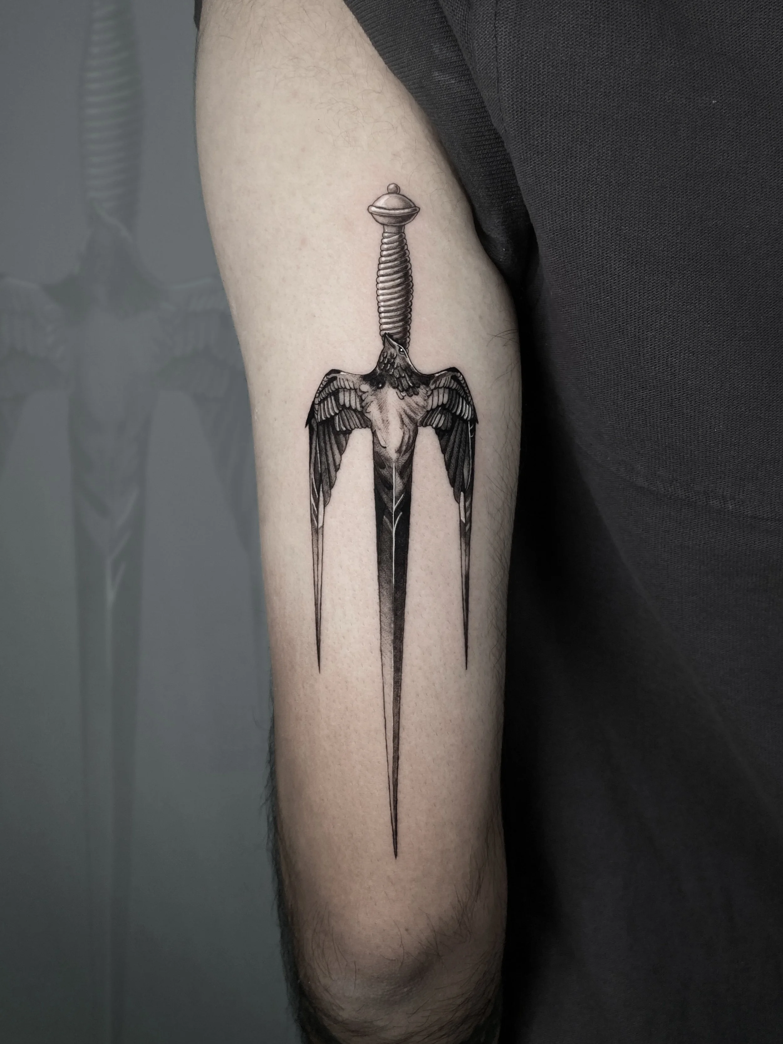 Black and gray tattoo of a sword with wings on the blade on a person's upper arm.