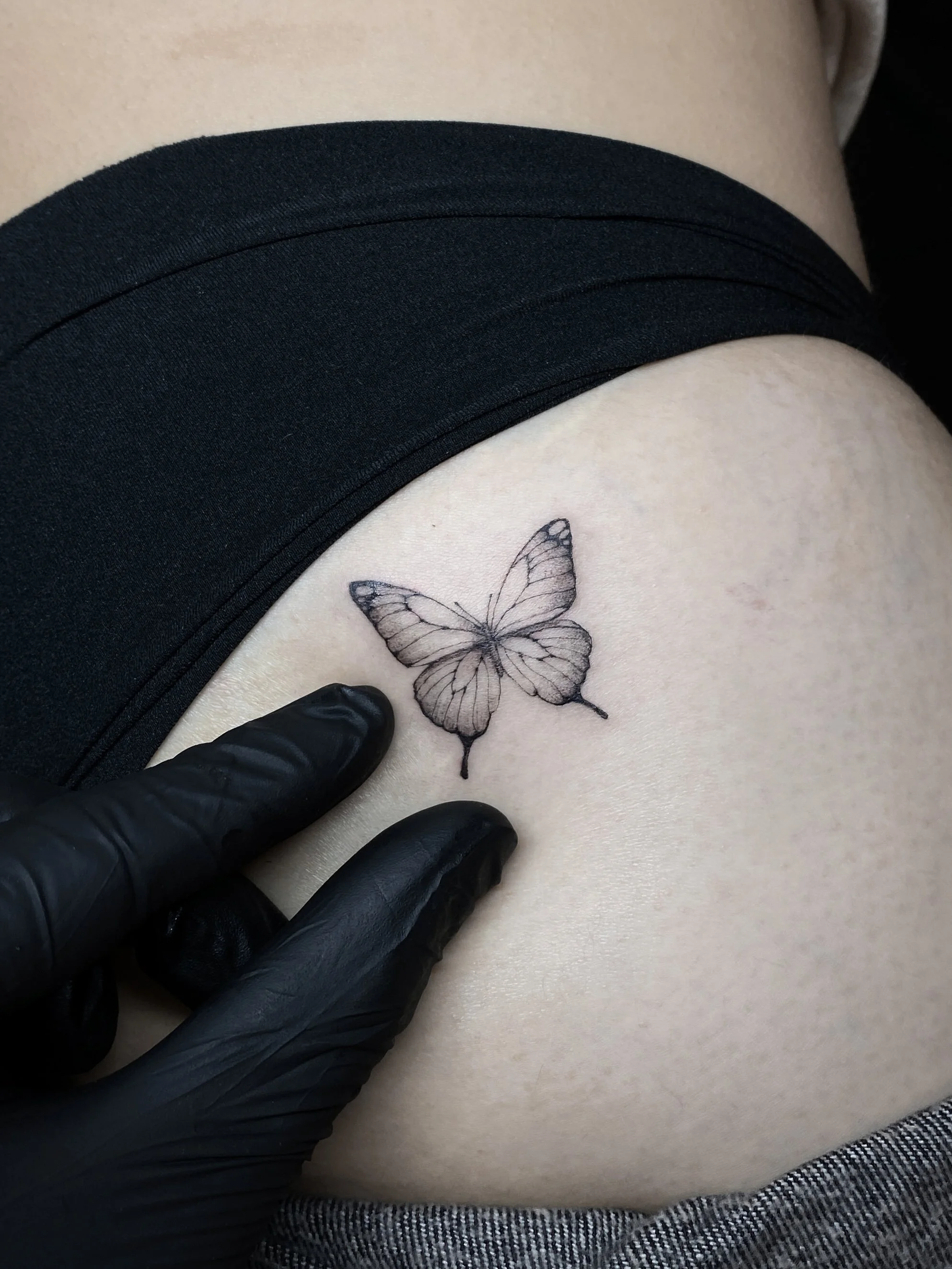 A close-up image of a tattoo of a butterfly on a person's skin, with someone wearing black gloves pointing at the butterfly. The person is wearing black shorts and gray clothing.
