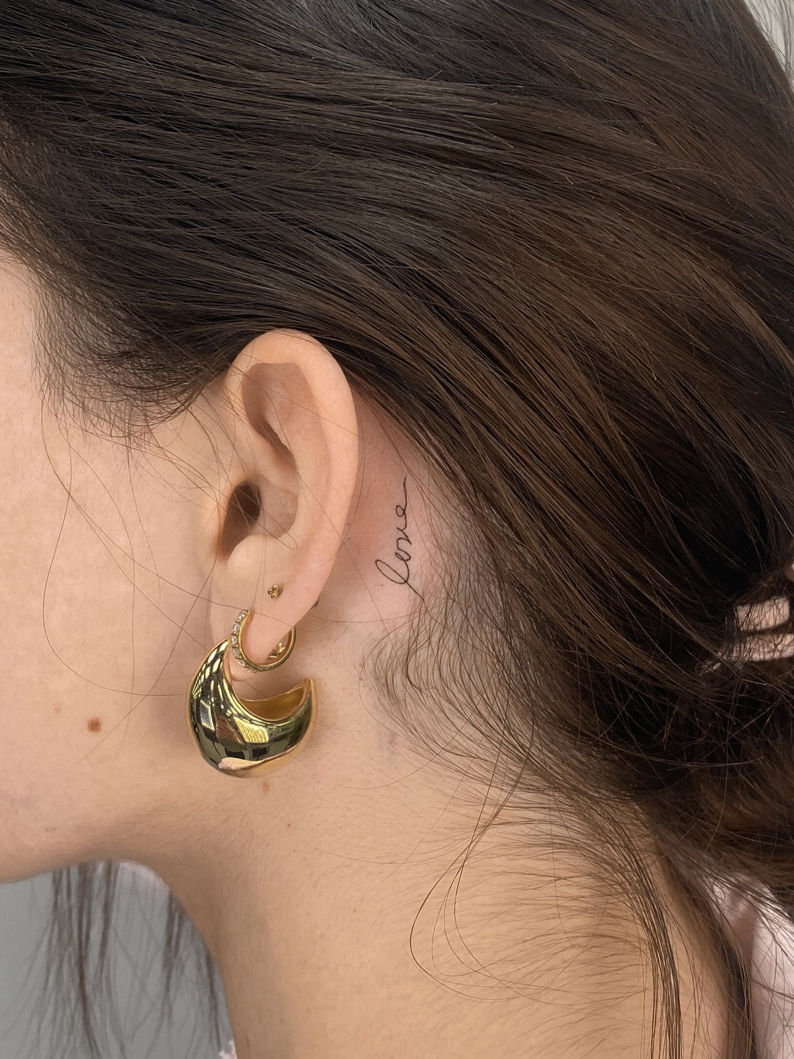 Close-up of a woman's left ear with multiple earrings, including a large gold hoop and a crescent-shaped gold earring, with a small tattoo saying "love" on her neck.