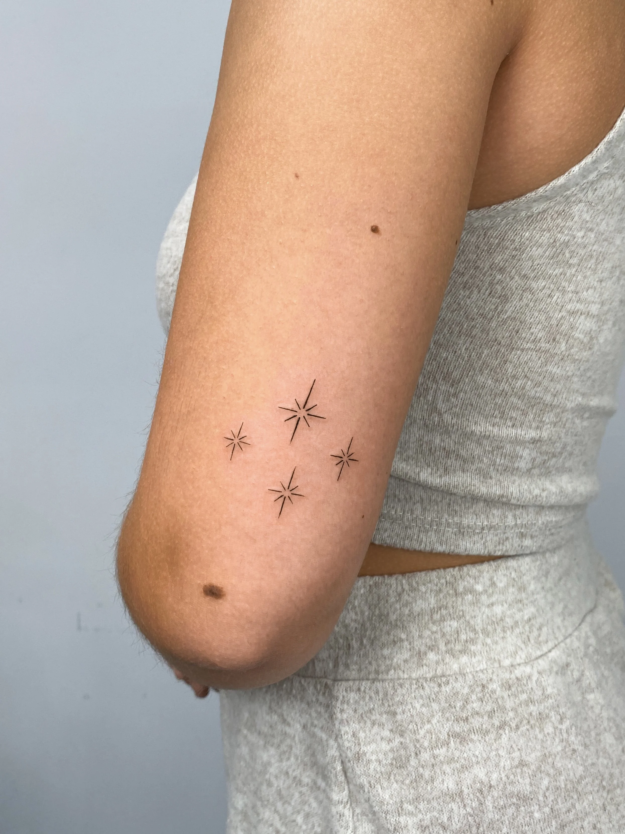 Close-up of a person's arm showing a minimalist tattoo of five small starbursts arranged in a slight curve on the inner forearm.