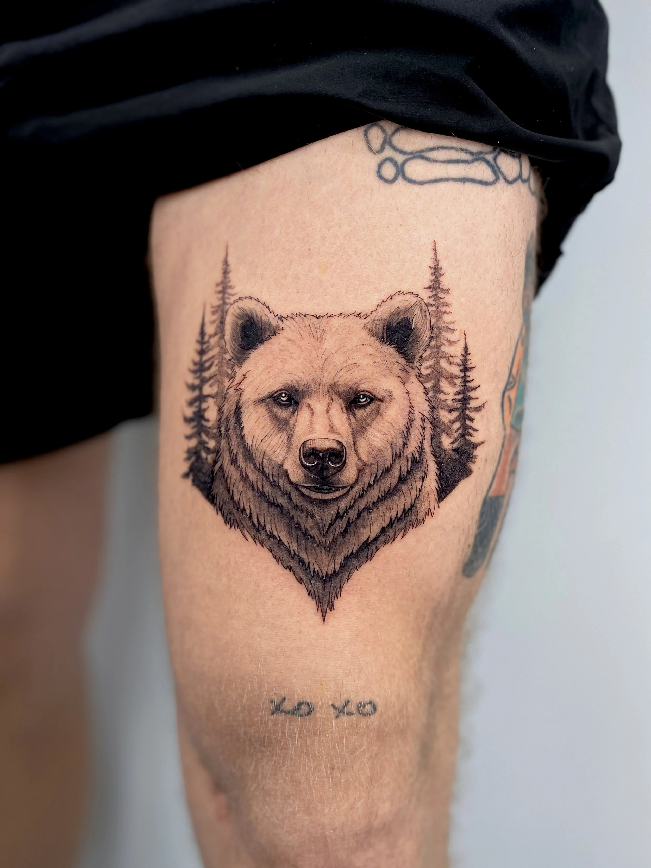 Tattoo of a bear's face with trees in background on a person's thigh, with a small tattoo below that reads 'xo xo'.