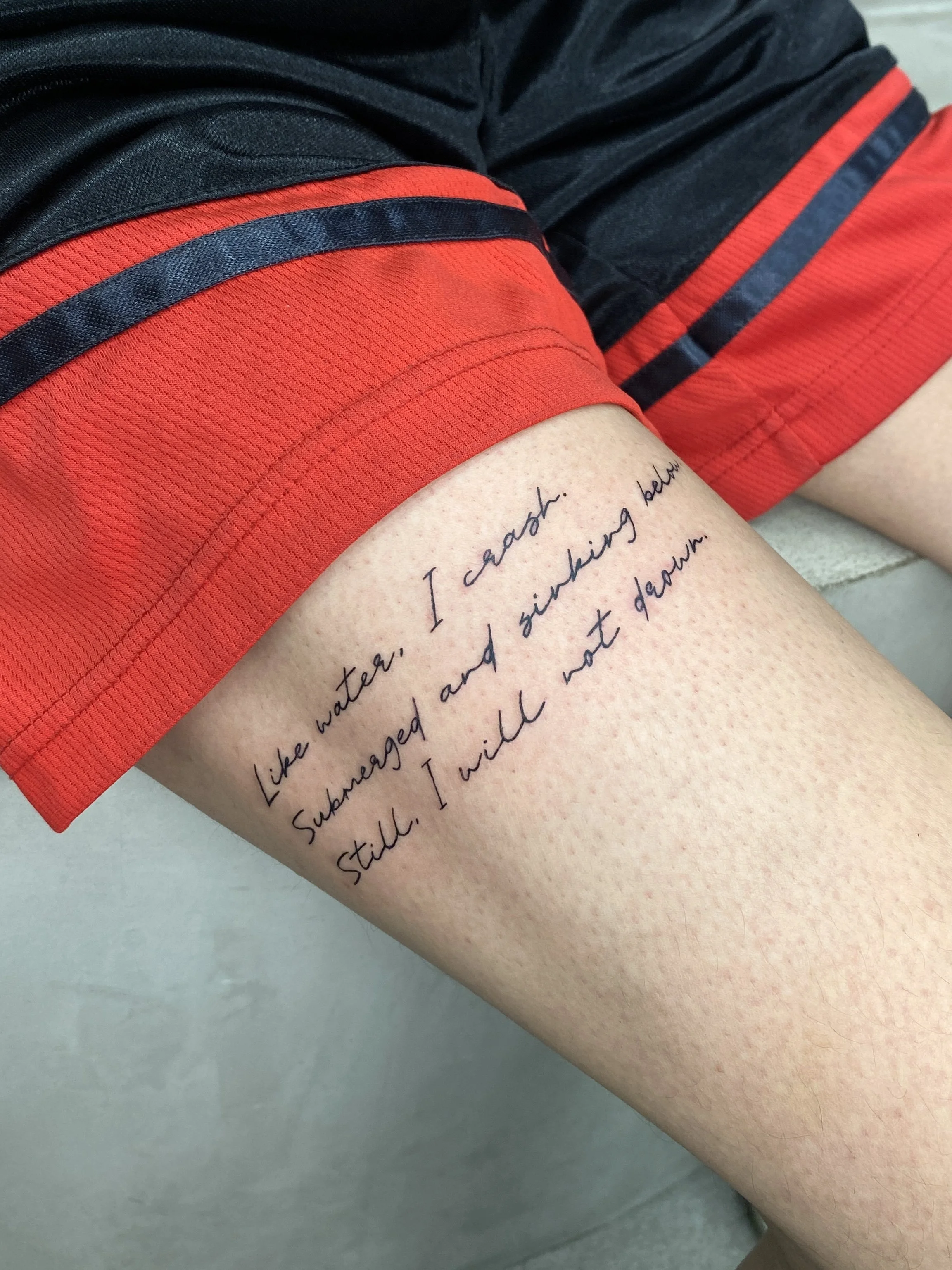 A person's thigh with a handwritten quote tattooed on it, visible on the skin, wearing black and red athletic shorts.