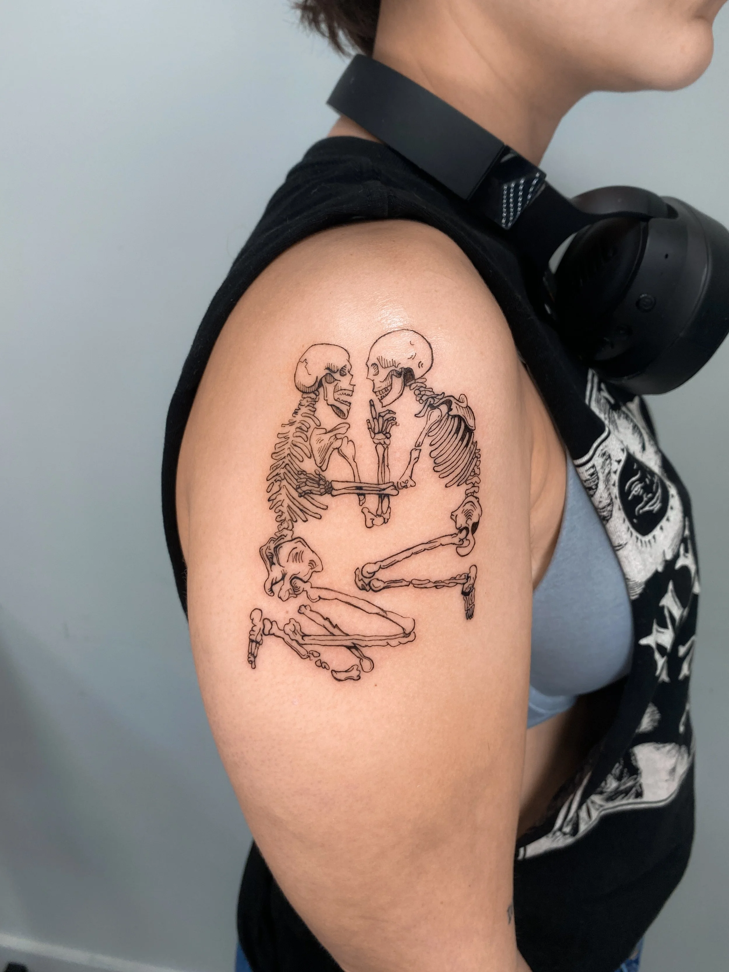 Tattoo of two skeletons facing each other, holding a small skeleton between them, on a person's shoulder.