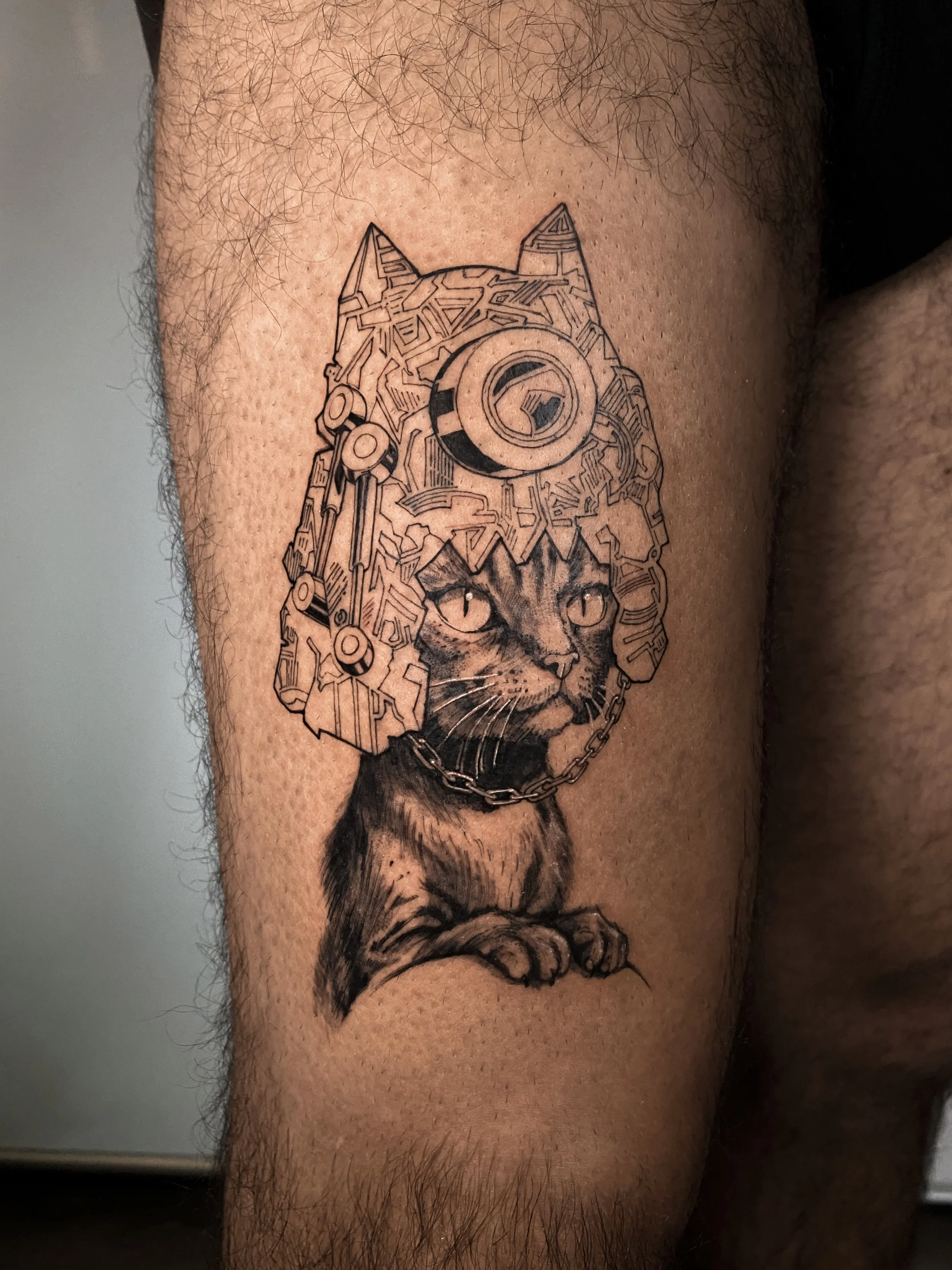 Tattoo of a cat wearing a cyborg-style helmet with circuitry and mechanical details, with a chain around its neck, on a person's thigh.