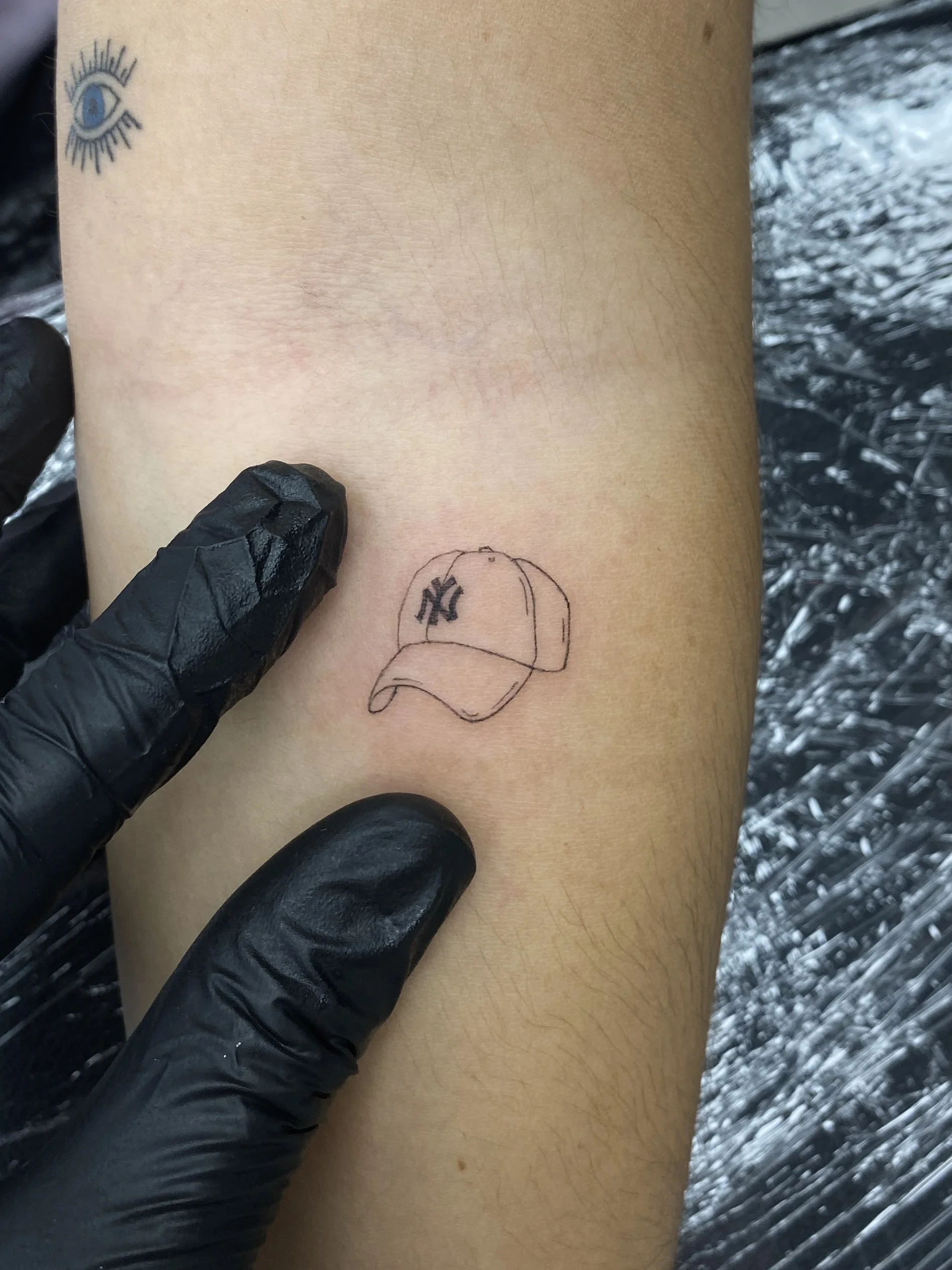 A tattoo artist's gloved hand is pointing at a small sketch of a baseball cap with the NY logo, on a person's skin.