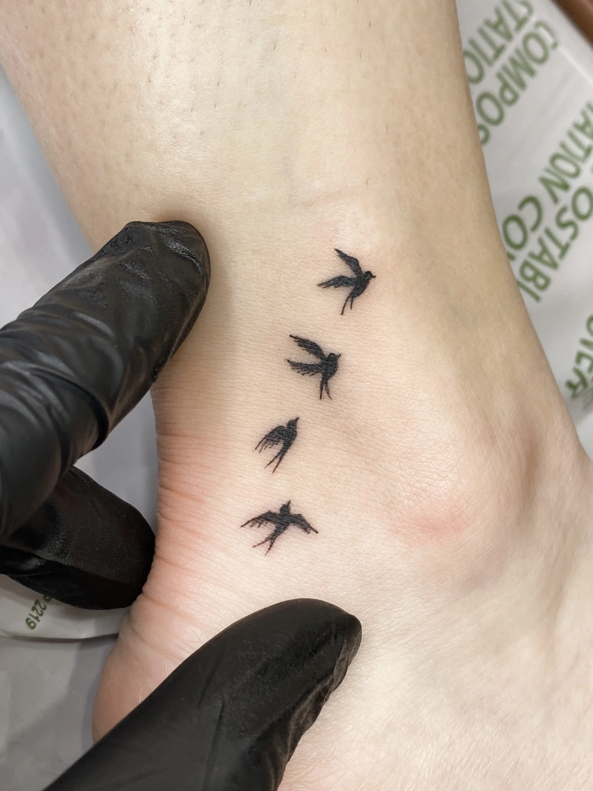 A fresh tattoo of four small black birds in flight on a person's wrist, with a gloved hand gently touching the tattoo.