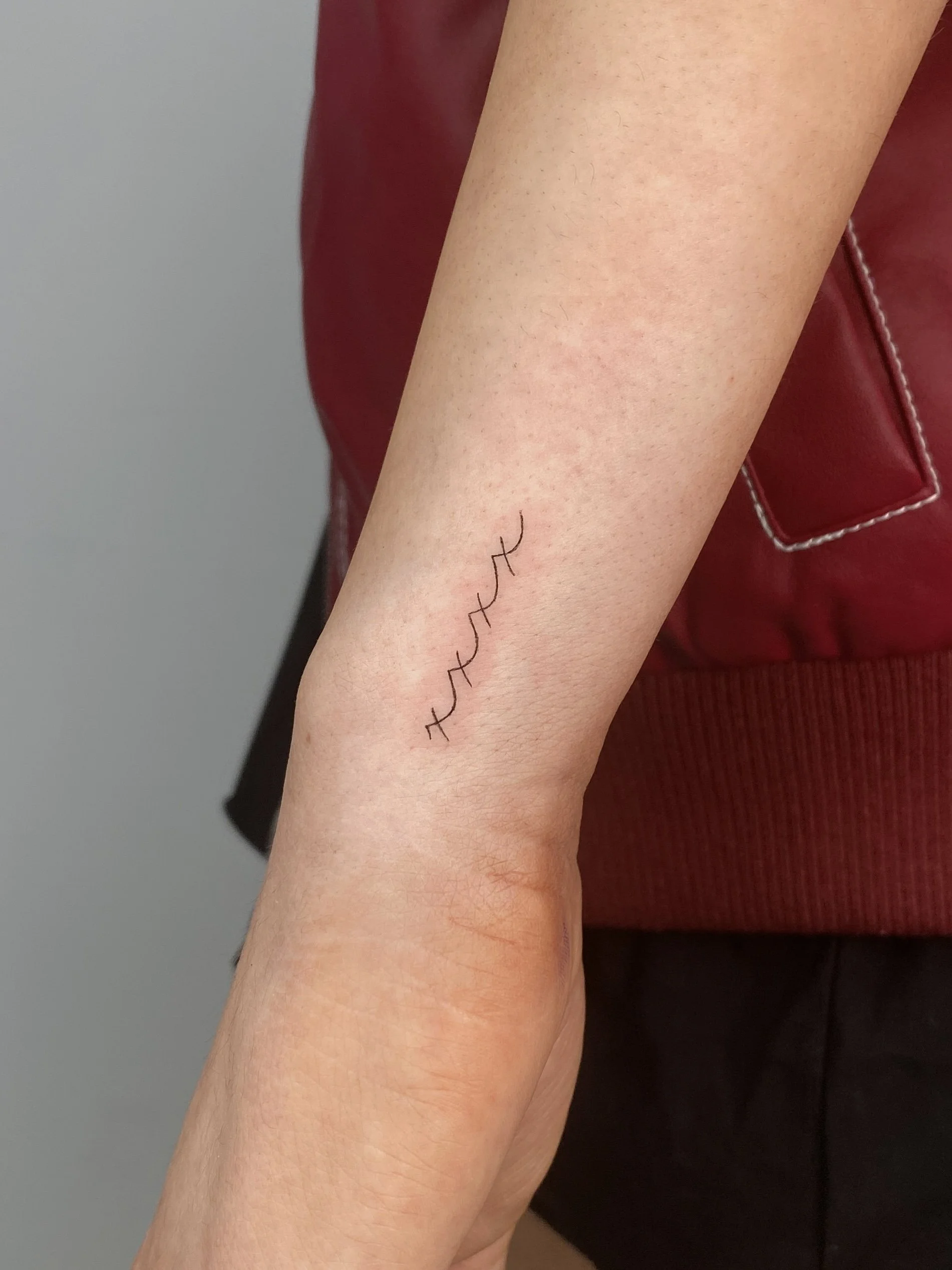 Close-up of a person's inner forearm with a small, black, minimalist tattoo of a heartbeat line.