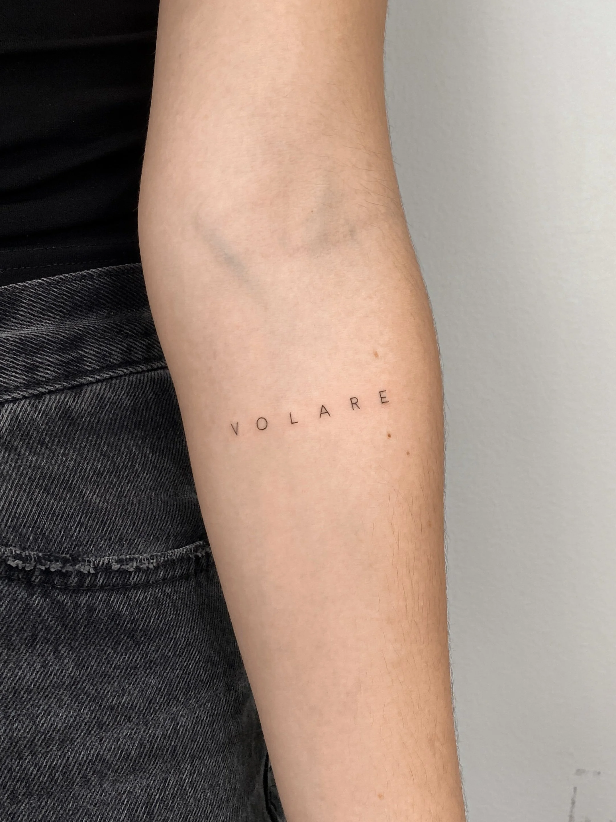 A tattoo of the word 'VOLARE' in minimalist black ink on a person's inner forearm.