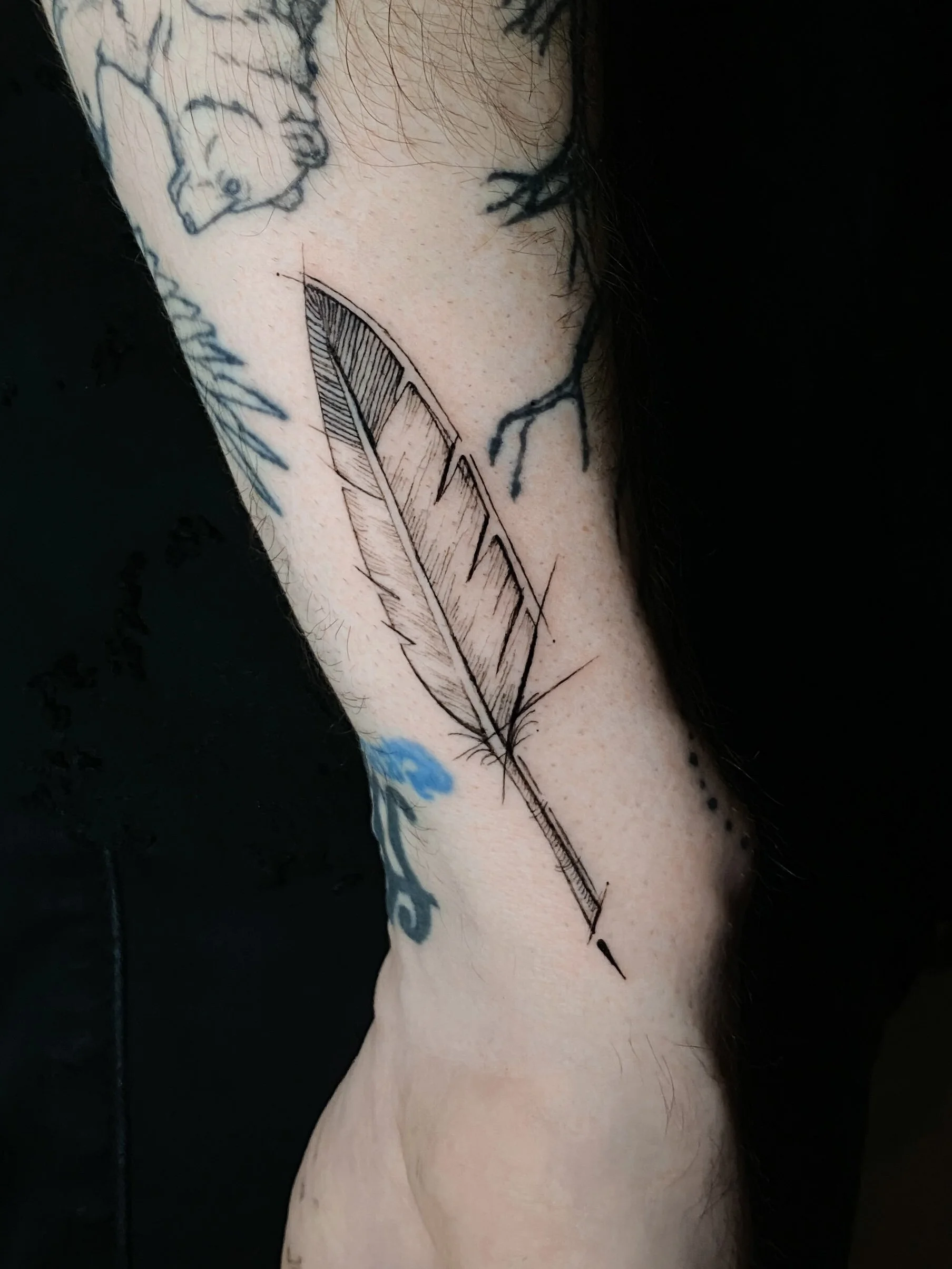 Black outline tattoo of a feather on a person's arm with other tattoos partially visible.