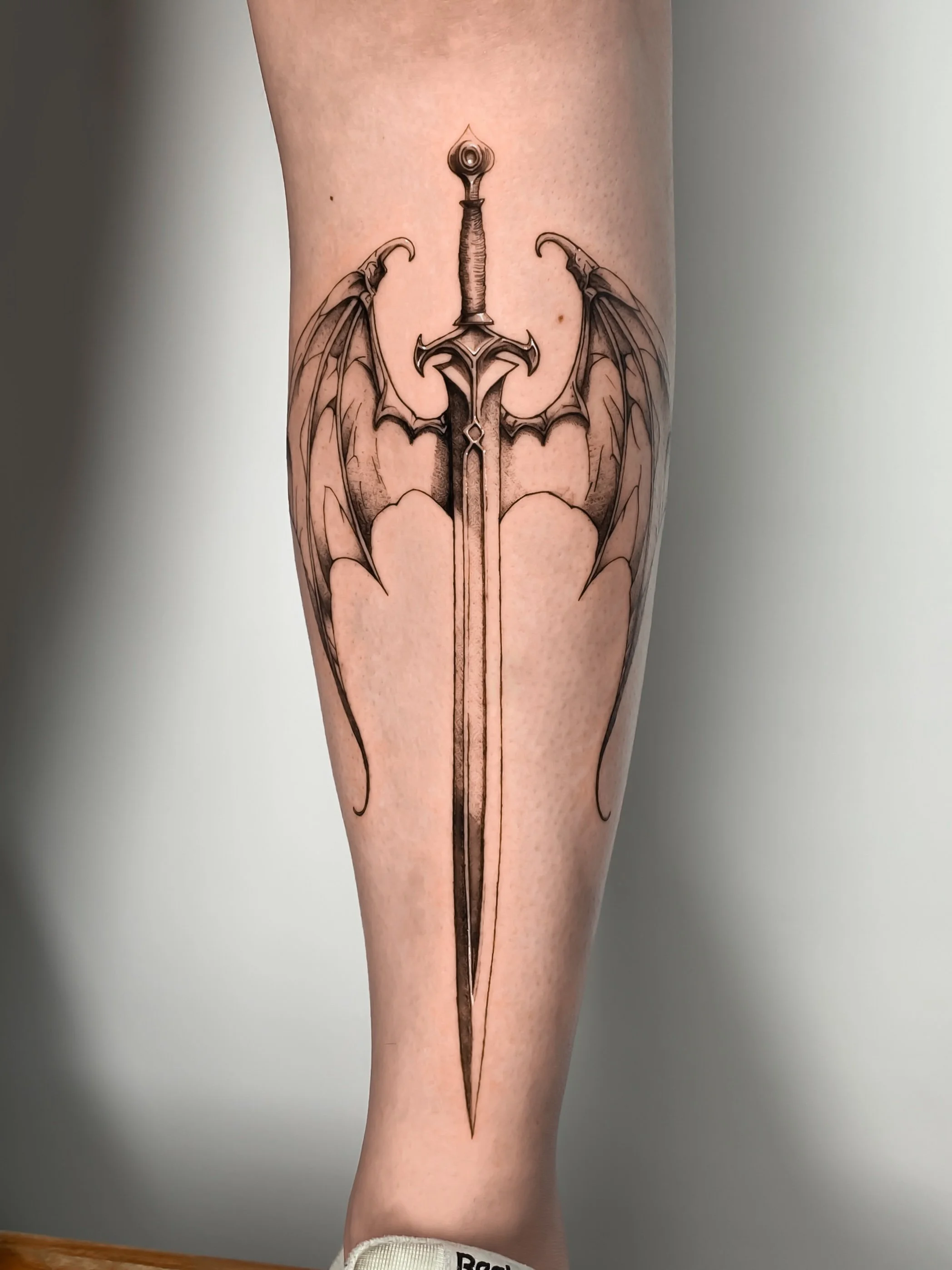 Tattoo of a sword with bat-like wings on a person's calf.