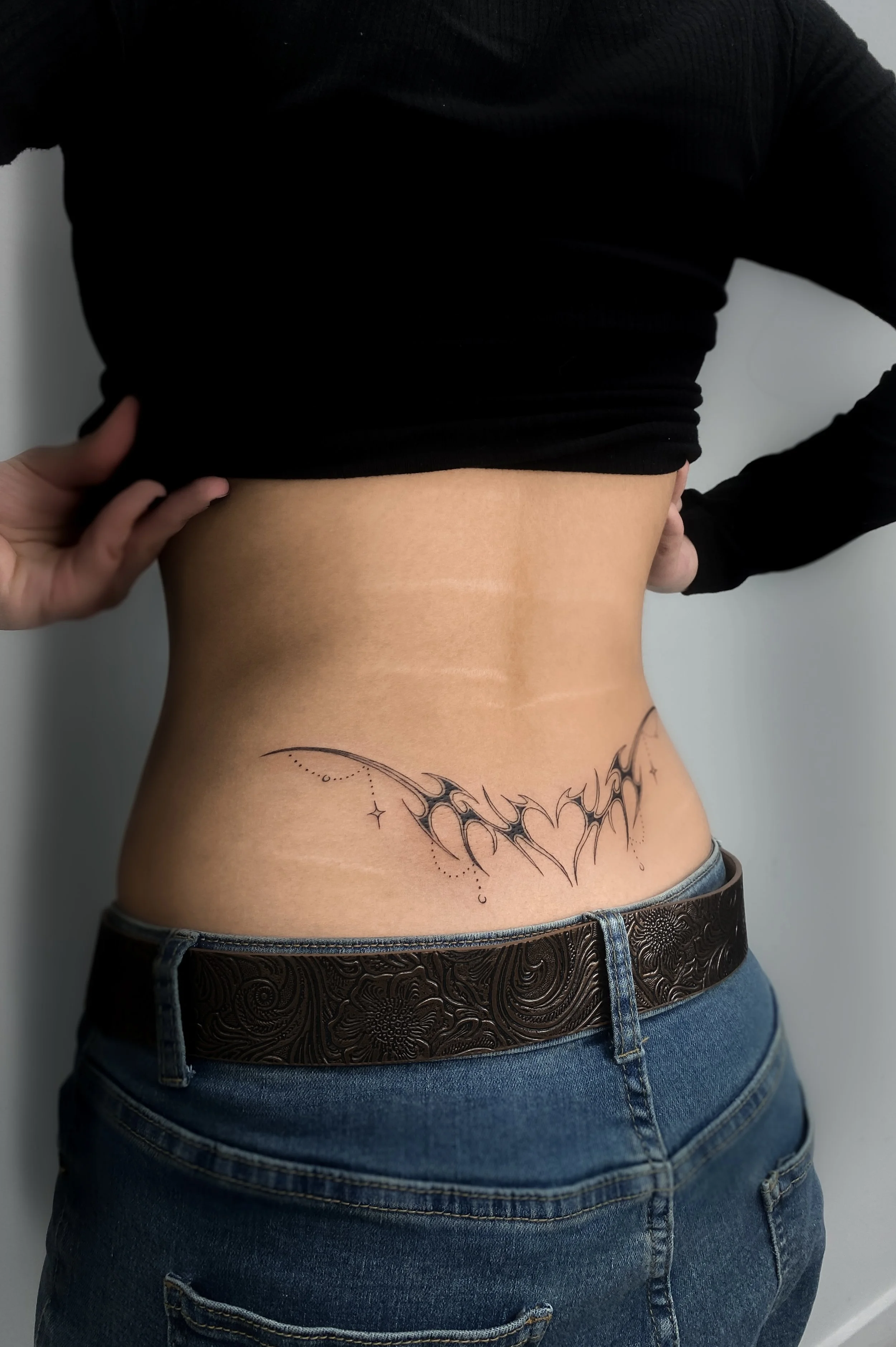 A person's midsection with a black bat-shaped tattoo on the lower abdomen, wearing blue jeans and a black top lifted to reveal the tattoo.