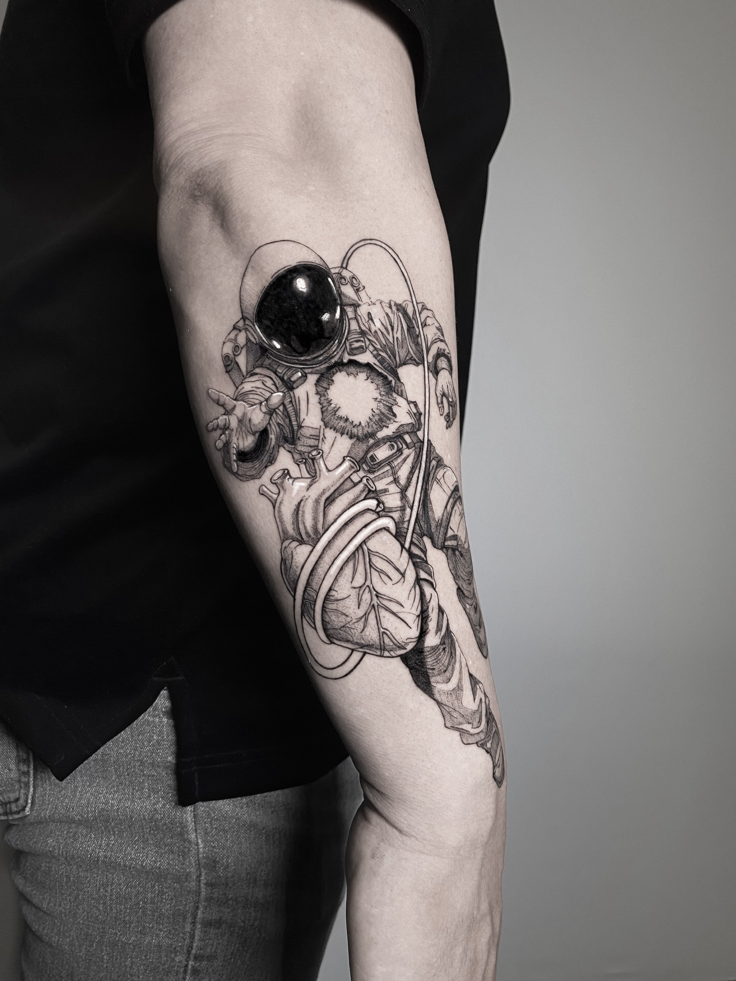 Black and gray tattoo of an astronaut reaching out with one hand, with a detailed human heart and tubing beneath.