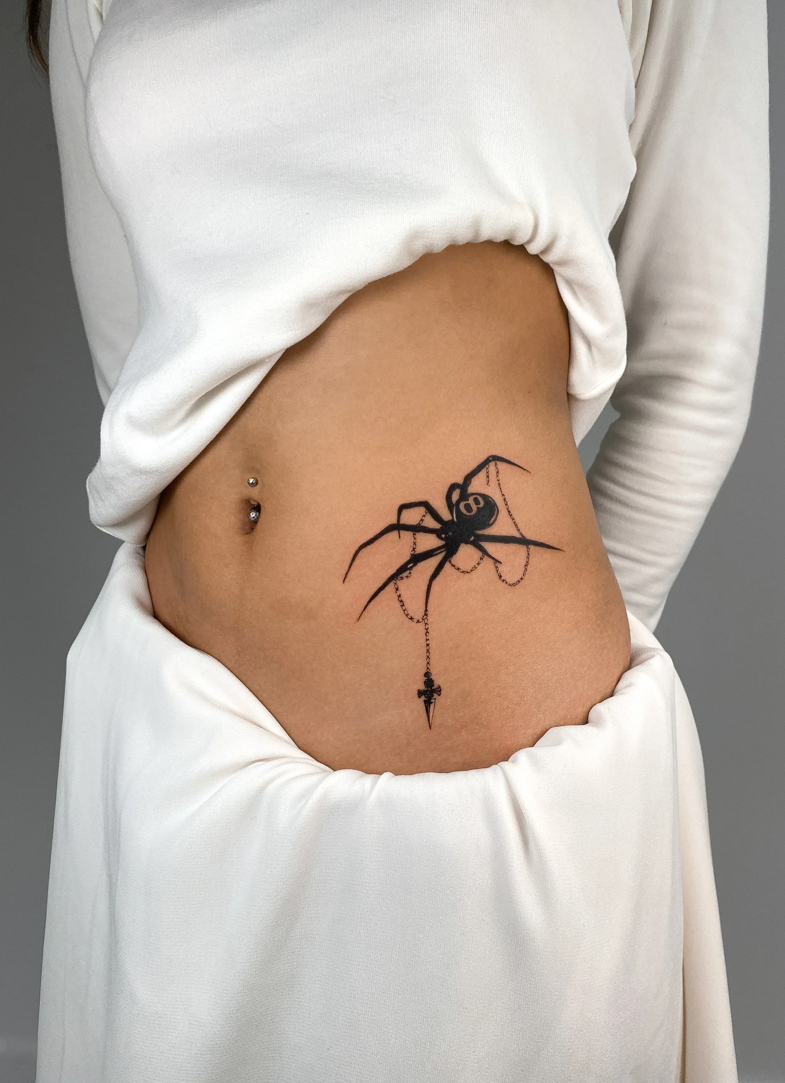A tattoo of a black spider holding a chain with a small dagger pendant on the person's side.