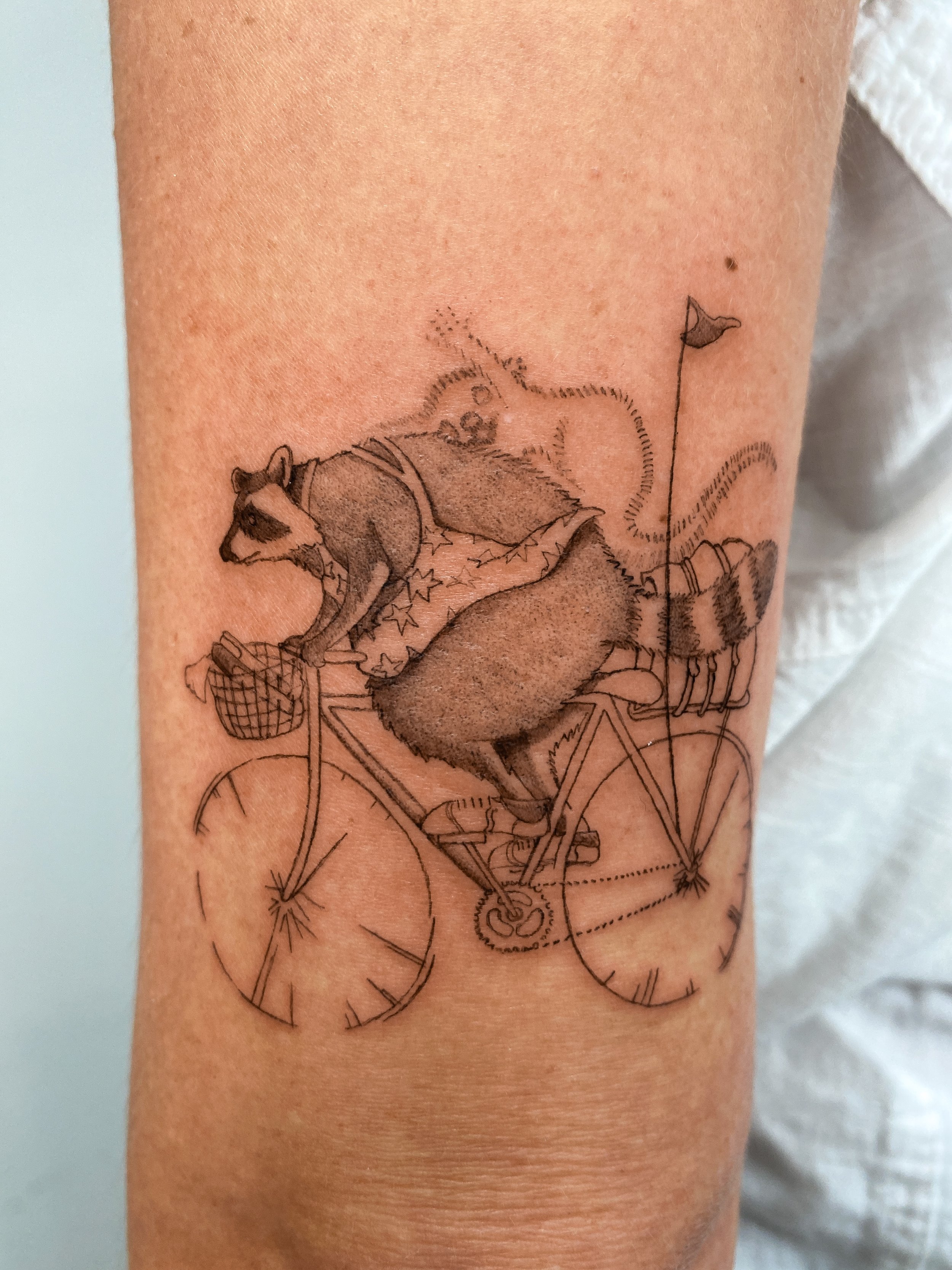Tattoo of a raccoon riding a bicycle, with a small raccoon on its back and a flag on the rear of the bike.
