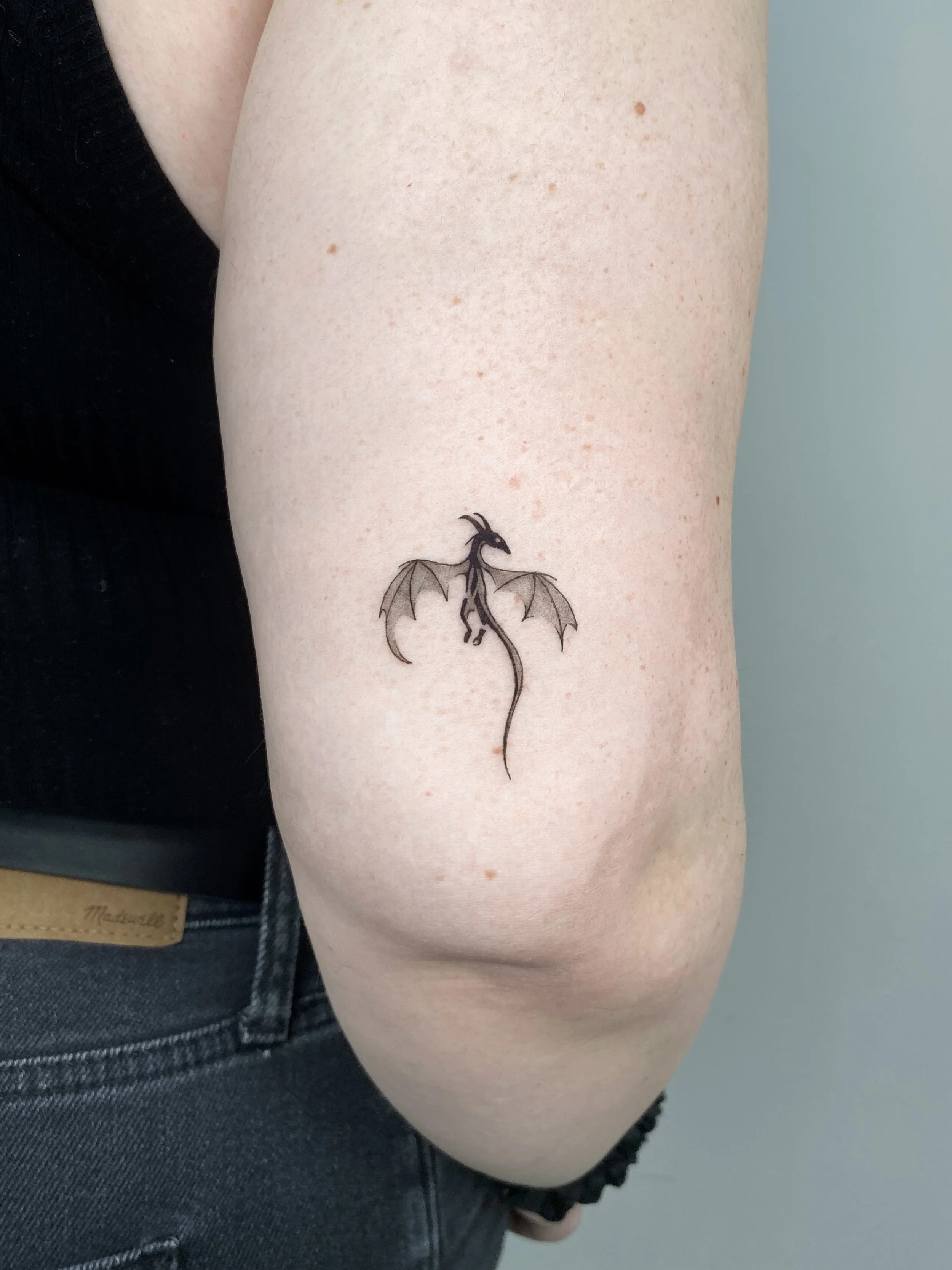 Close-up of a person's upper arm with a small tattoo of a black dragon with wings, a long tail, and an angular head.