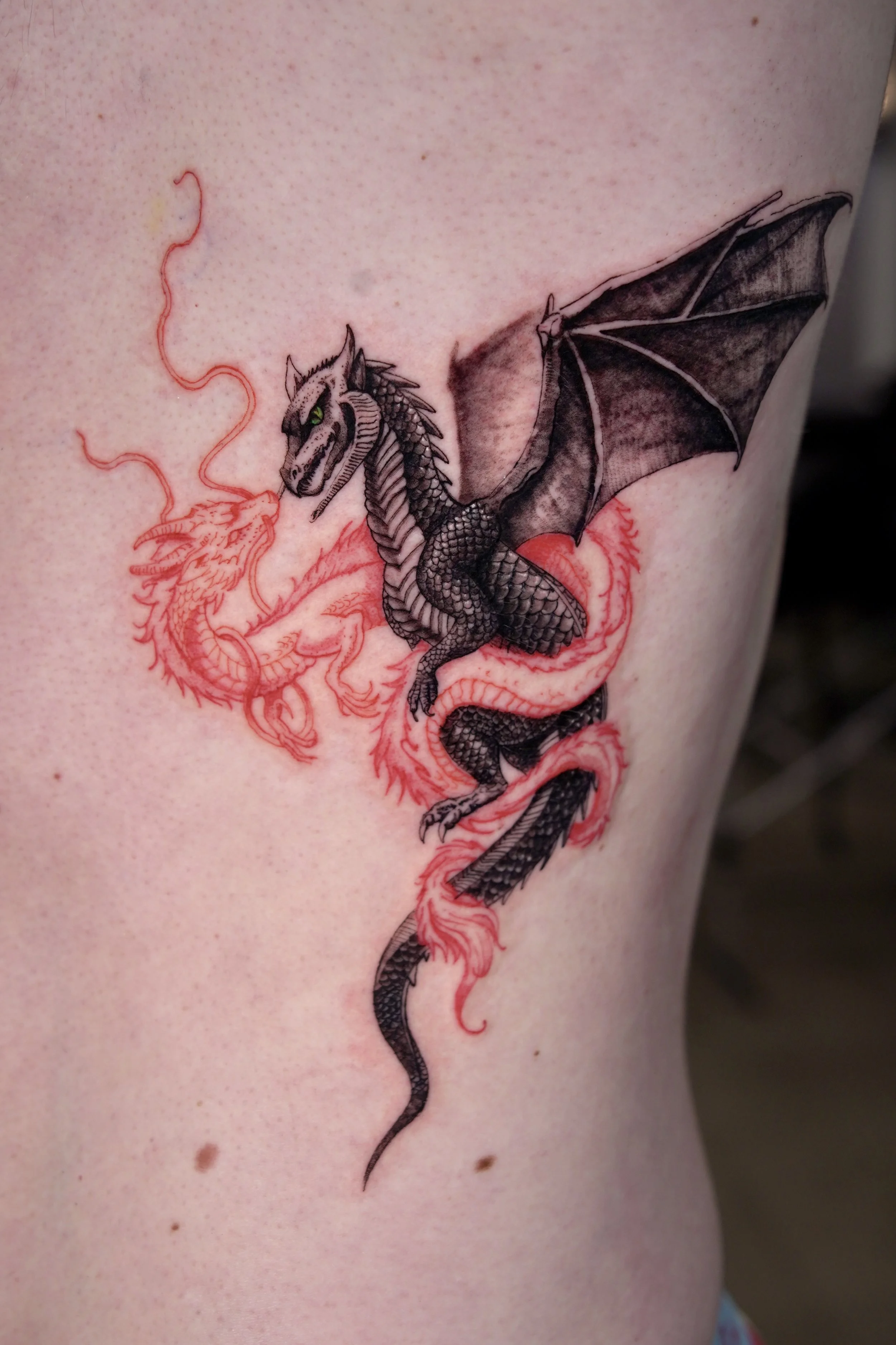 A tattoo of a black and gray dragon with green eyes, breathing fire. The dragon's body is detailed with scales, wings, and claws. There is a red outline of a different, simpler dragon in the background.