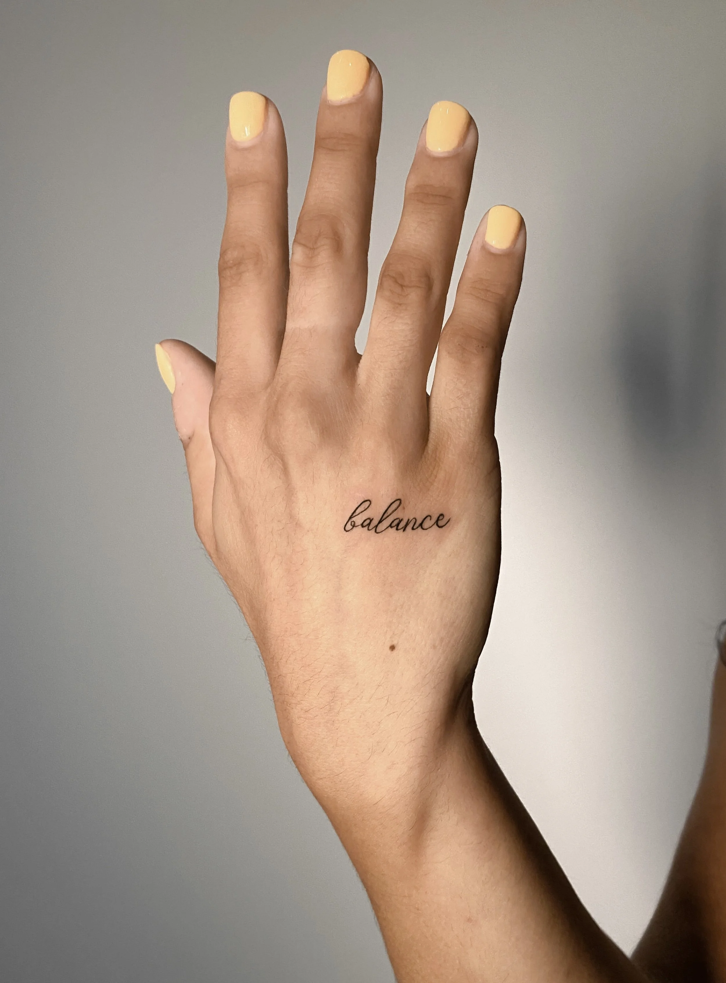 A hand with yellow-painted nails showing the word balance tattooed on the wrist against a plain background.