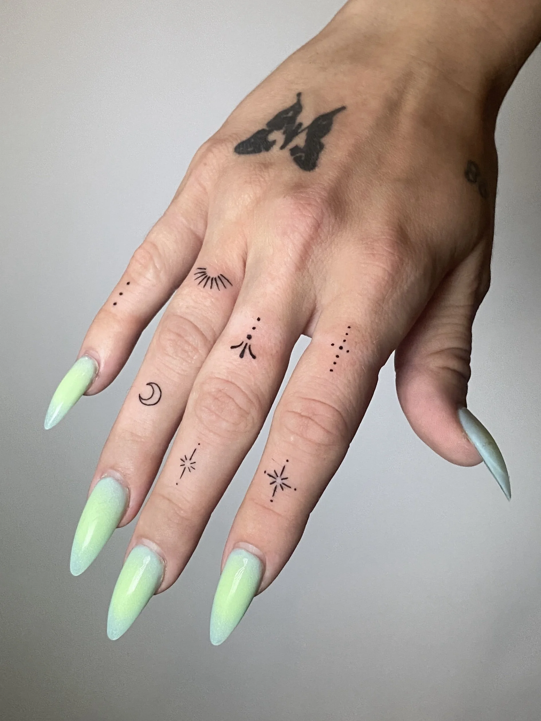 A hand with long, pointed nails painted in a light green color. The hand has several small tattoos, including a flying bird on the wrist and various small symbols on the fingers, such as dots, a crescent moon, and starbursts.