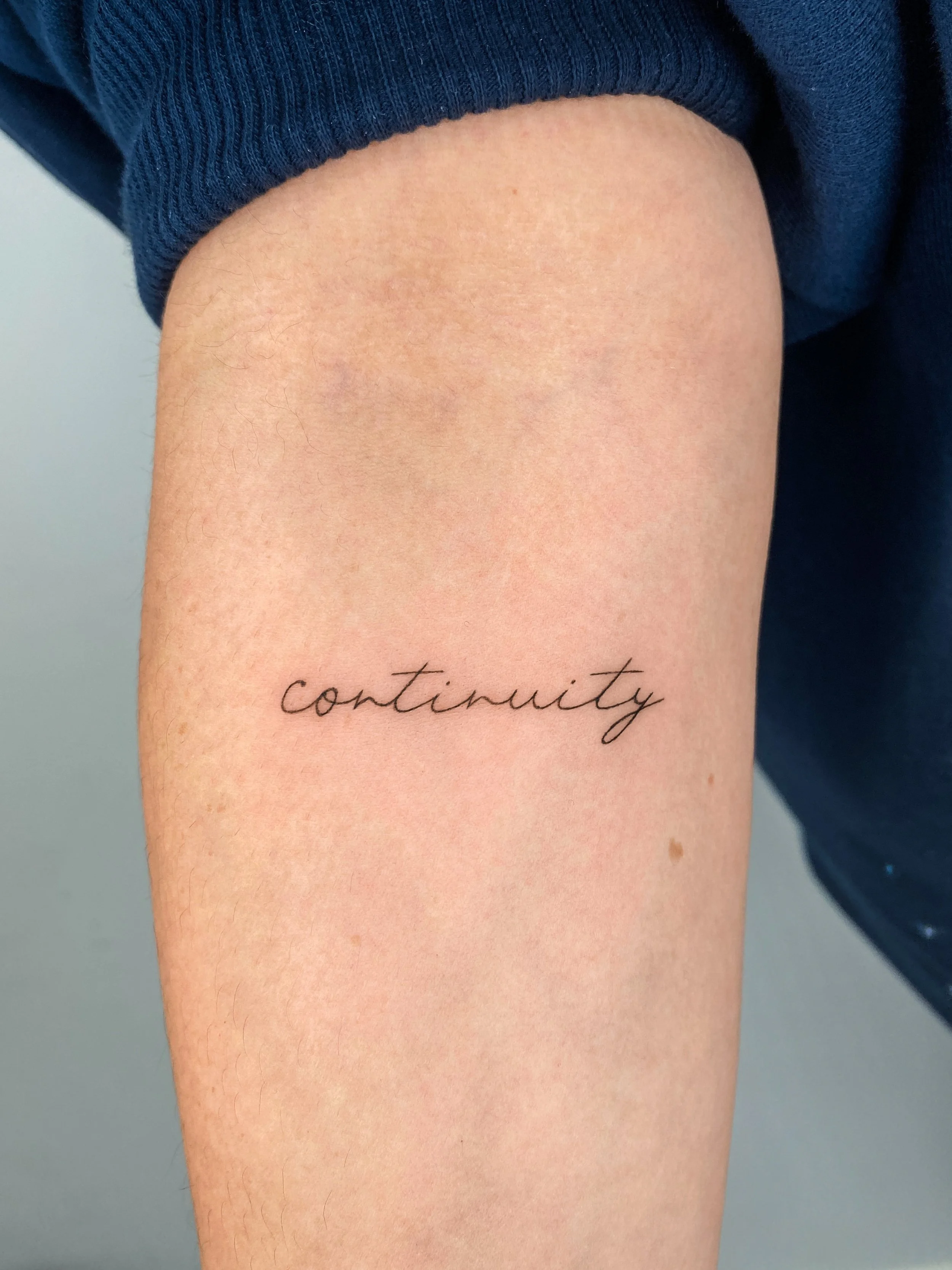 Tattoo of the word 'continuity' in cursive on a person's upper arm.